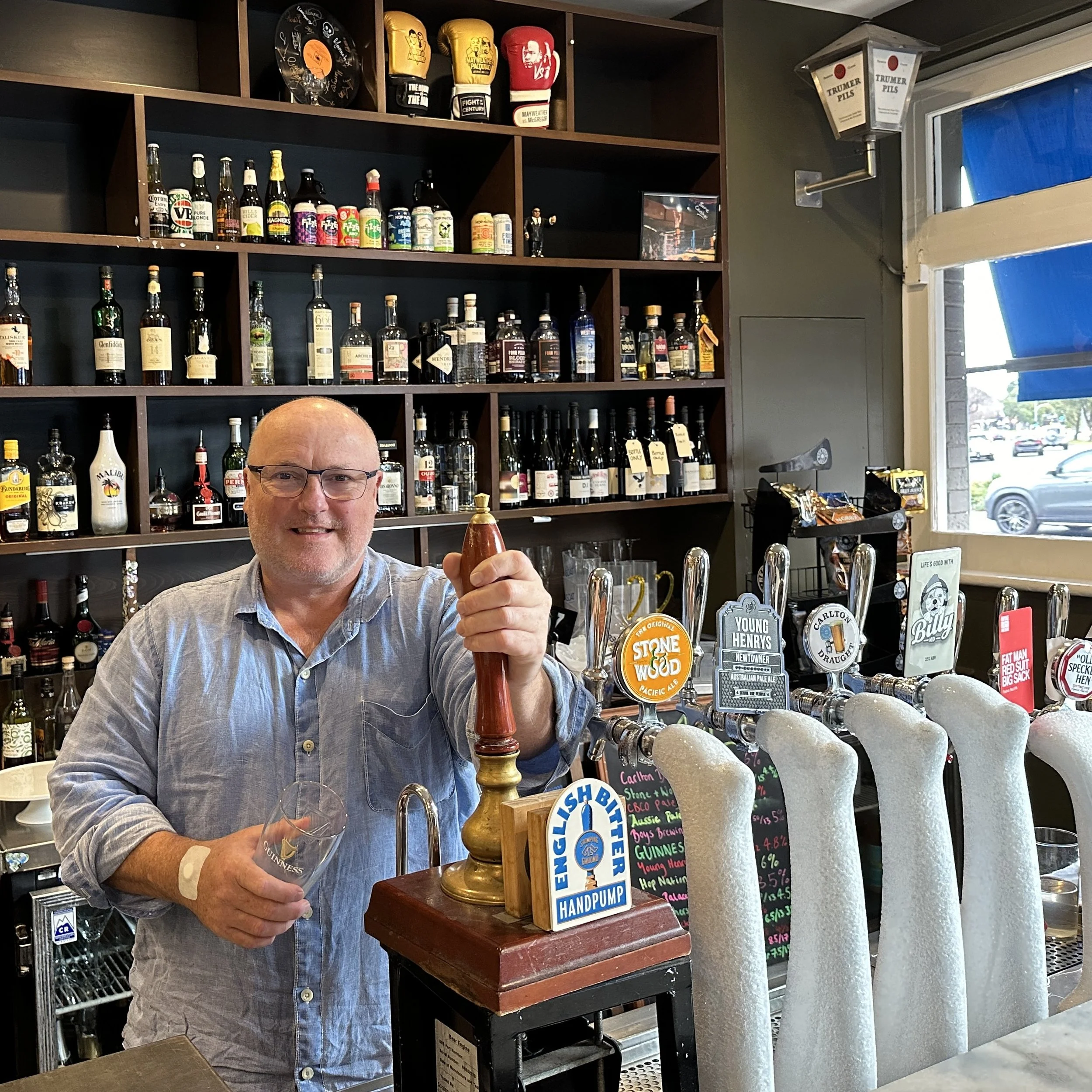 A smiling man with glasses and a grey shirt standing at a bar, holding a wooden hand pump with a tap and a branded glass, in a pub with shelves of liquor bottles and beer taps behind him.