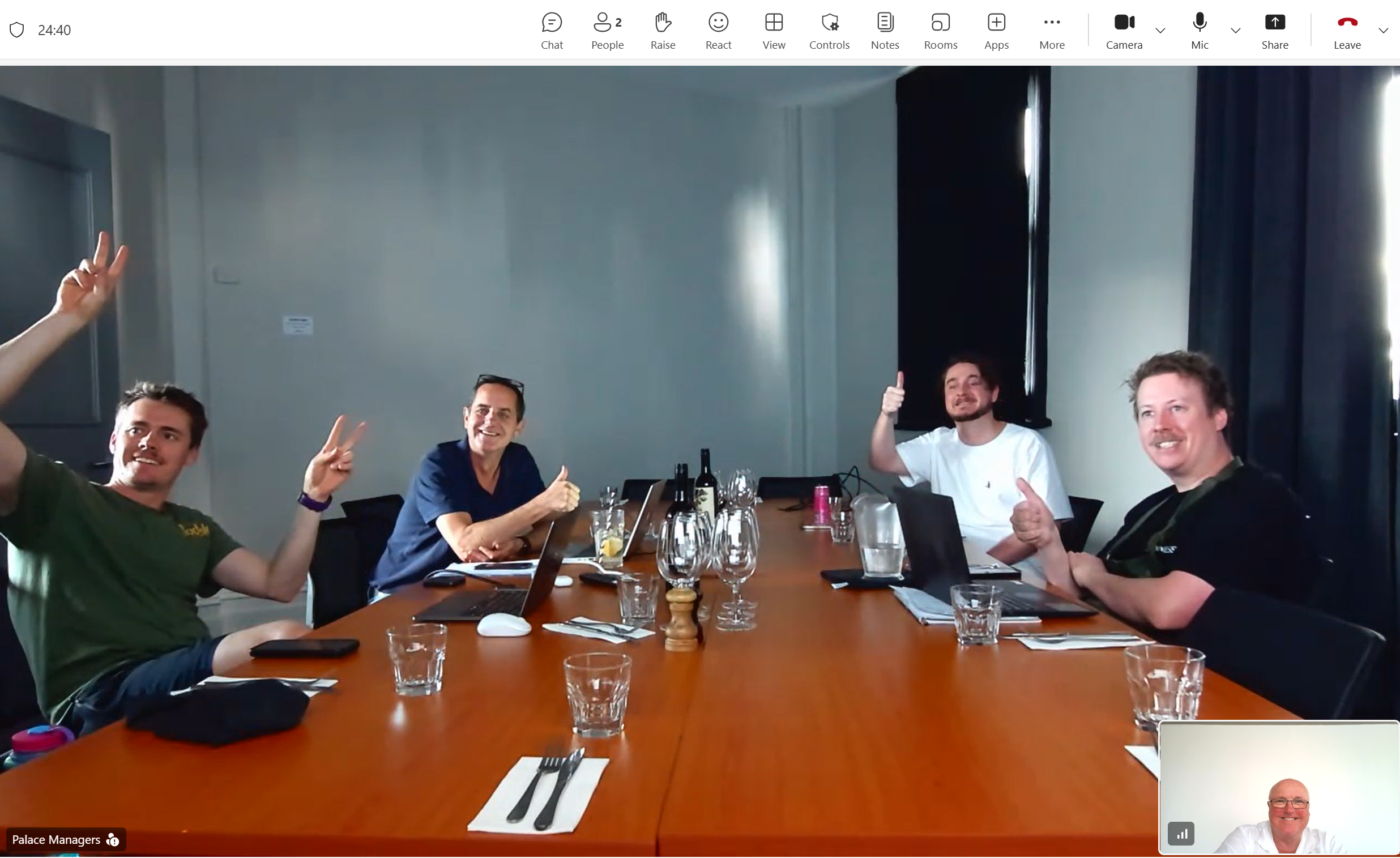 Four men sitting around a conference table smiling and making peace signs or thumbs up gestures during a meeting in a bright room.