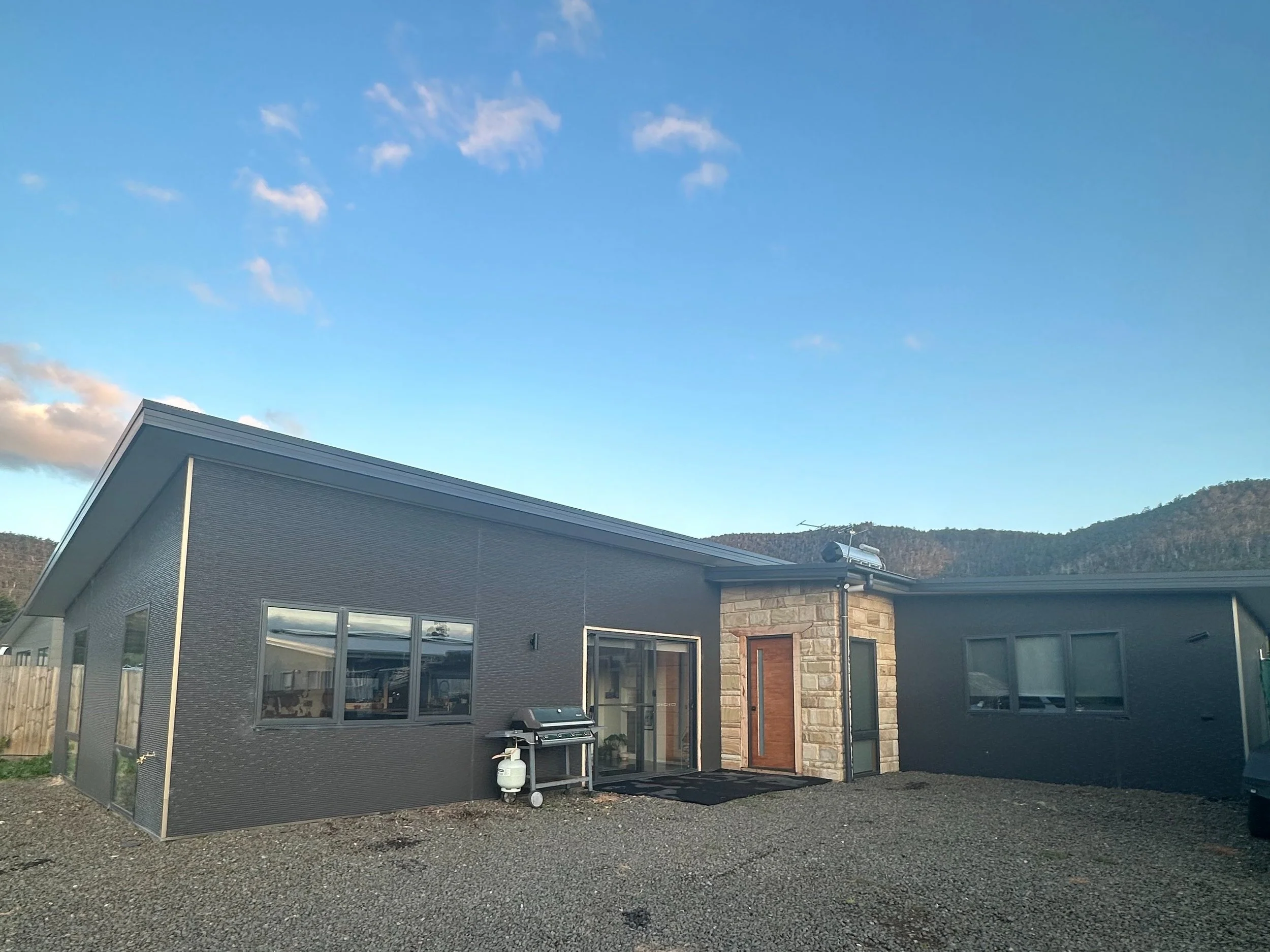 Modern house with gray exterior, stone accents at the entrance, and a barbecue grill outside on gravel ground, under a blue sky with some clouds.