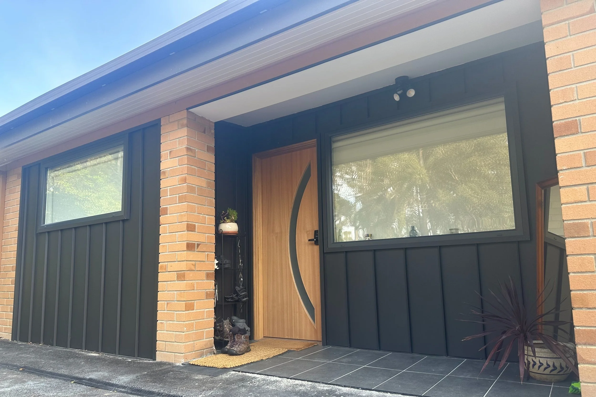 Modern house entrance with black siding, brick pillars, and a wooden door with a curved glass window. There is a large window to the right and a smaller window to the left. Decorative plants and shoes are near the door.