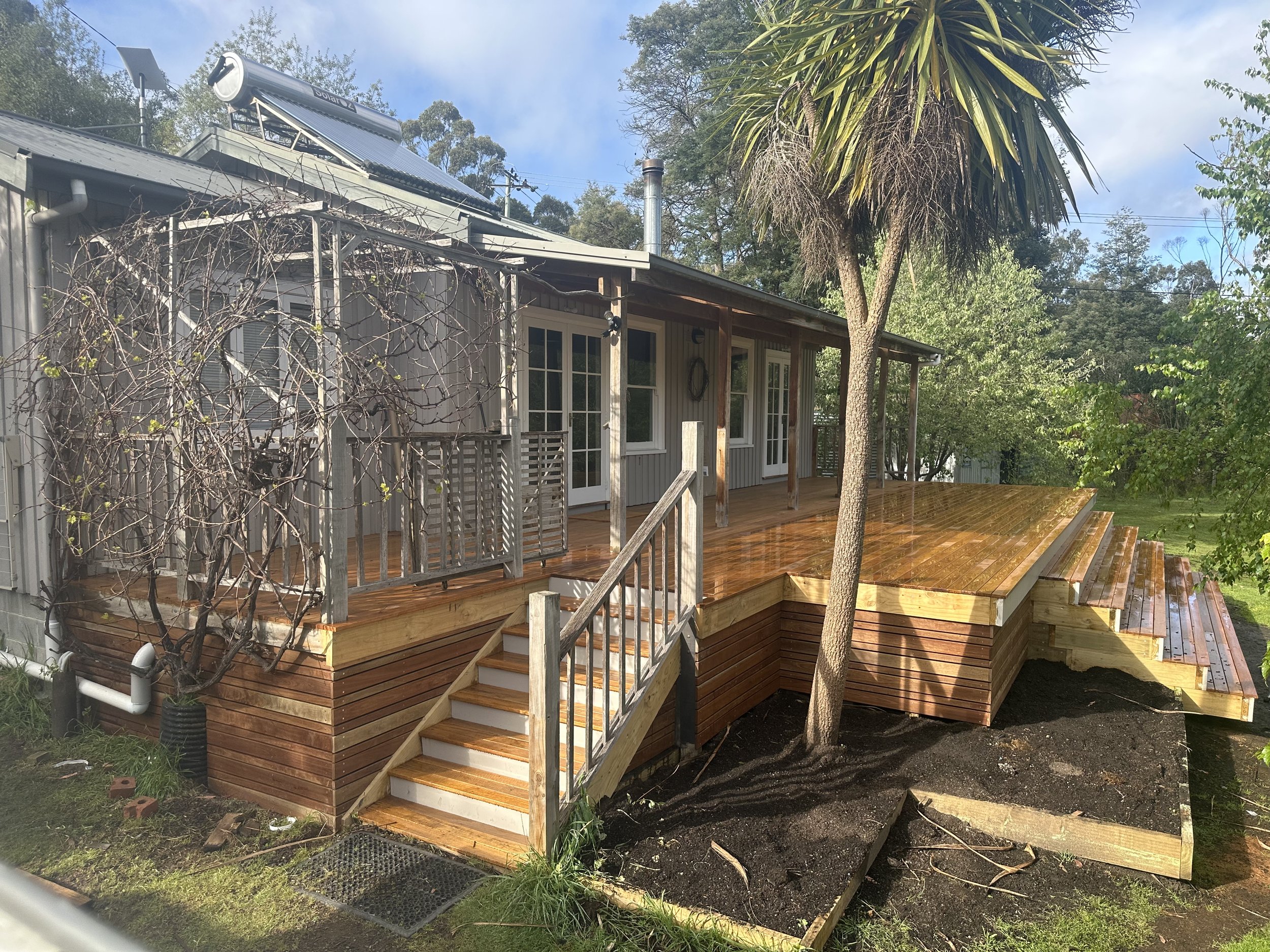 A newly built wooden deck attached to a house with white siding. The deck has stairs leading down to the yard, some plants, and a tree nearby. The house has large glass doors and windows, and there is a solar water heater on the roof.