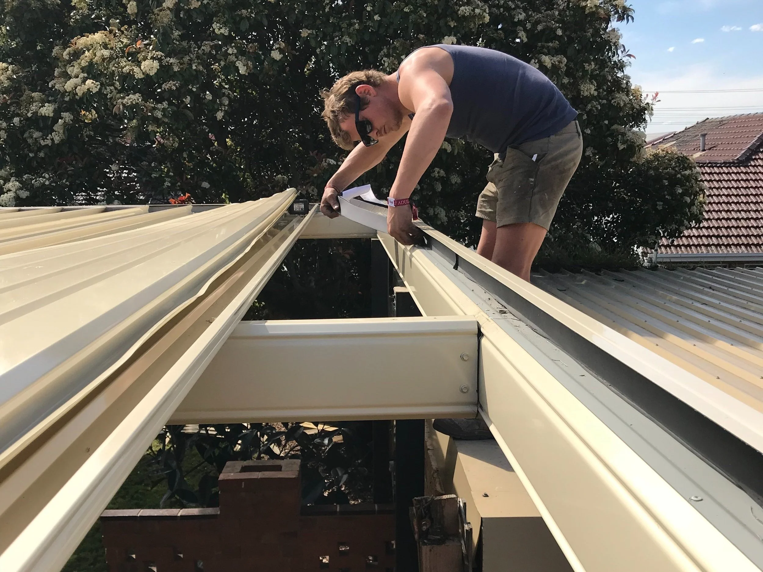 A man working on a roof, installing or repairing gutters, with greenery and houses in the background on a sunny day.