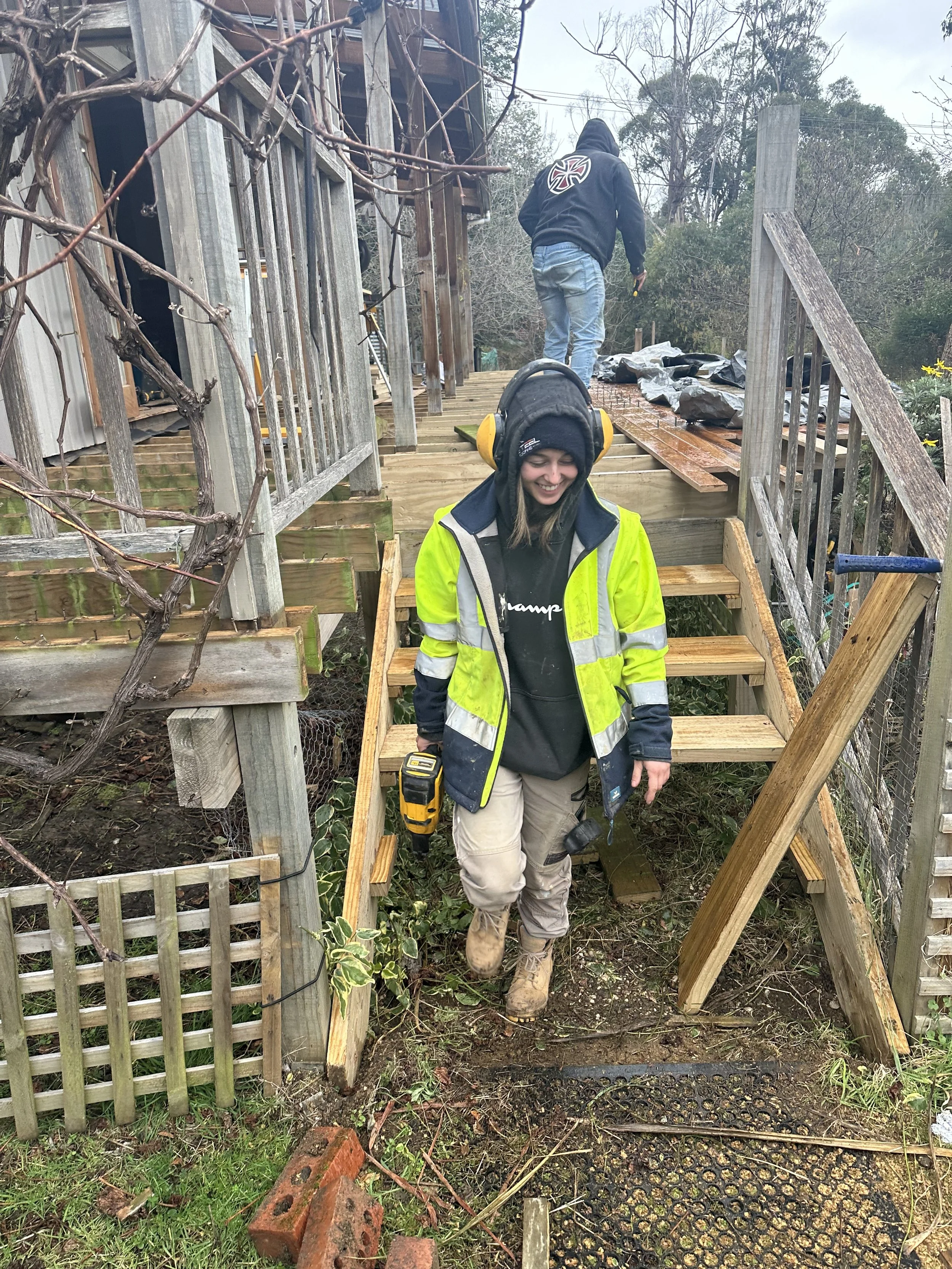 Women working on constructing wooden stairs on an outdoor deck, wearing safety gear and using cordless tools, with two men in the background.