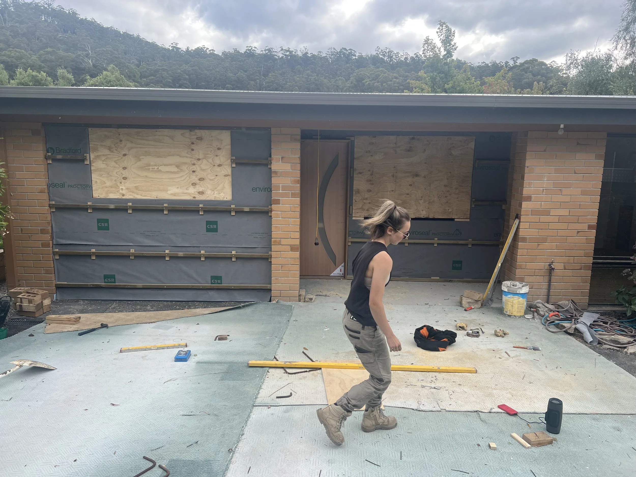 Woman wearing black sleeveless top, gray pants, and tan work boots on a construction site, working on the exterior of a house with brick walls and the garage door opening boarded up with plywood, surrounded by tools, materials, and construction debris.
