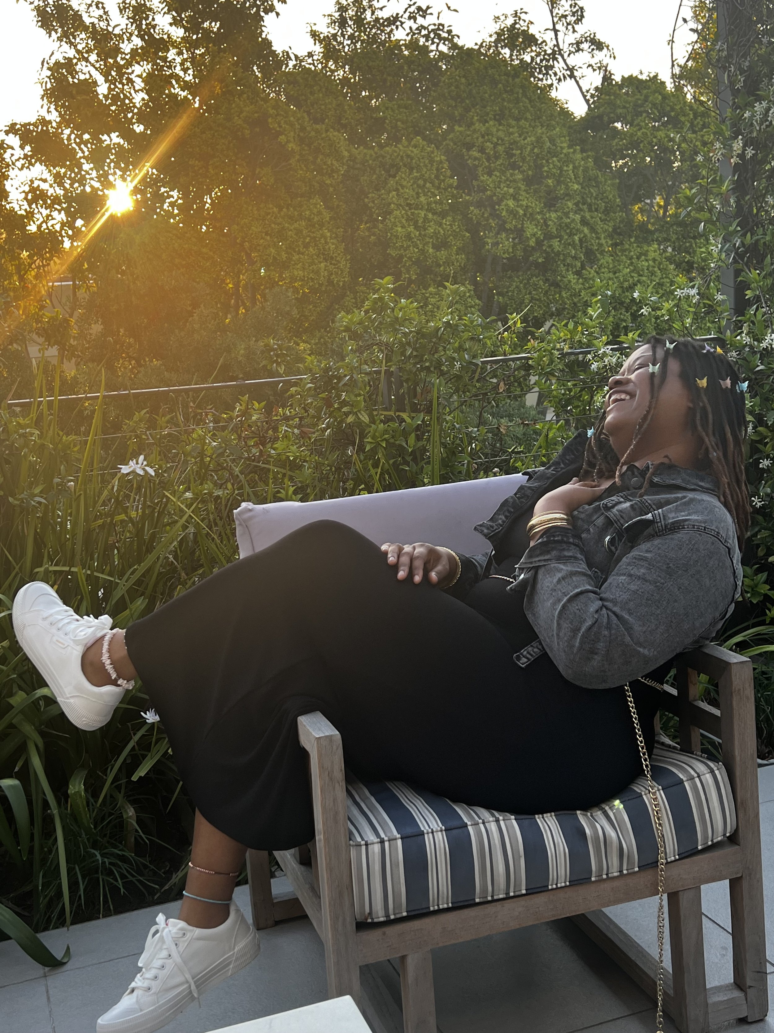 A woman with dreadlocks and butterfly hair clips is relaxing on a striped cushion on a wooden bench outdoors, surrounded by greenery and trees, during sunset. Swank sitting outdoors at sunset surrounded by trees and greenery.