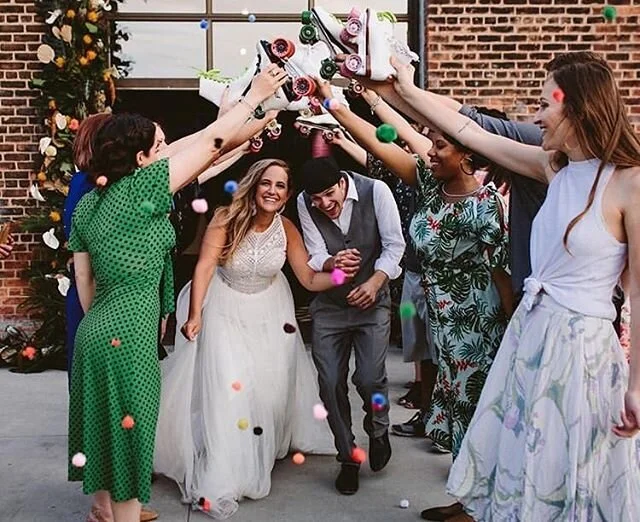 People celebrating a wedding with a confetti toss, including a bride in a white wedding dress and a groom in a gray vest and tie, surrounded by friends in colorful dresses, outside a brick building.