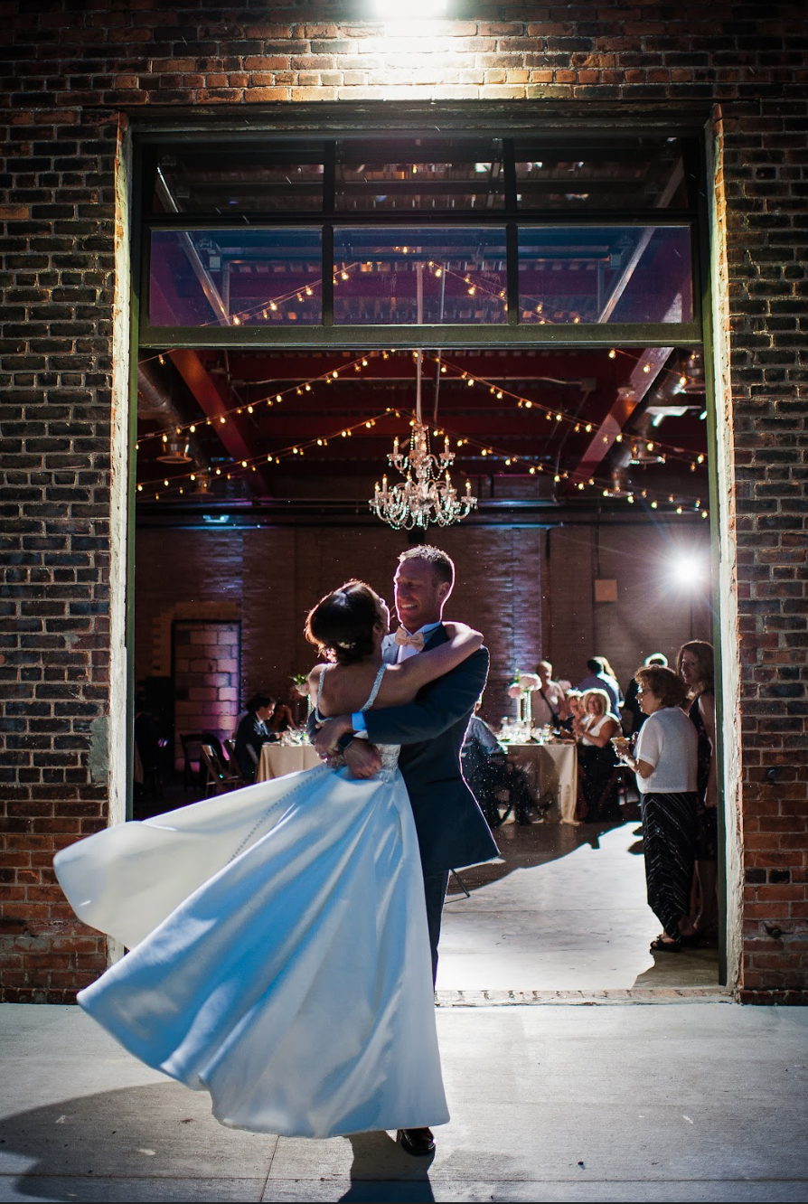 A wedding couple dancing inside a decorated venue, seen through a large brick archway. The bride is in a white dress, and the groom is in a dark suit. Guests are seated at tables and standing in the background, with festive string lights and a chande