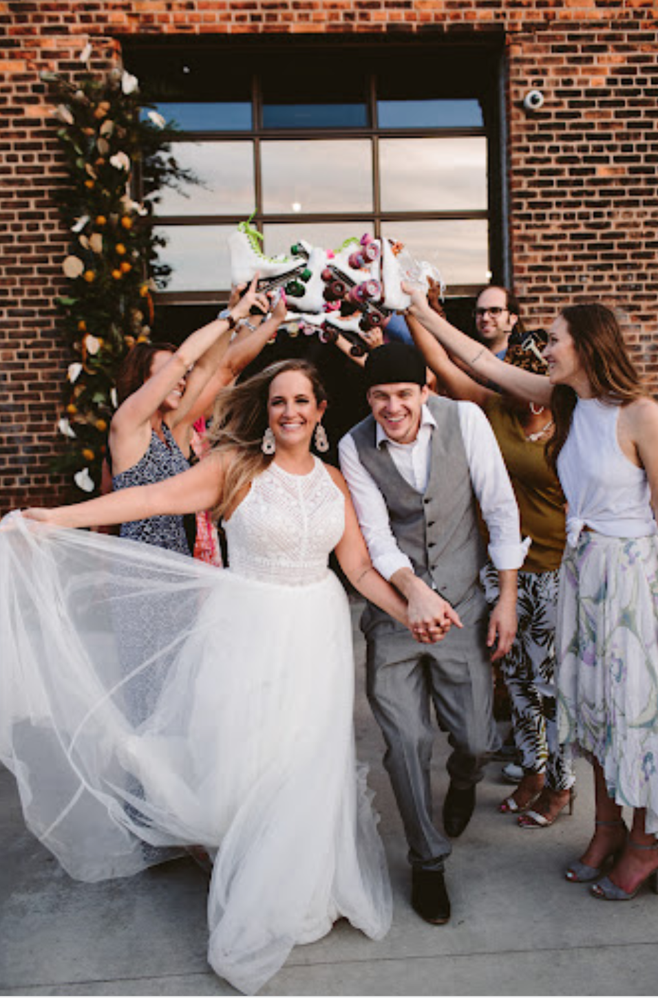 A joyful group of friends, including a bride and groom, celebrating with a cake topper of roller skates outside a brick building during daytime.