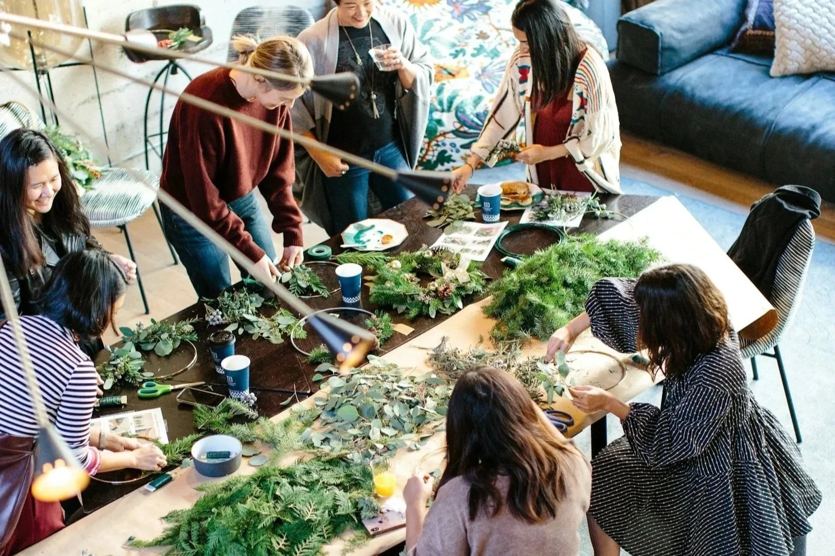 People gathered around a table making holiday wreaths with green foliage and pine branches.