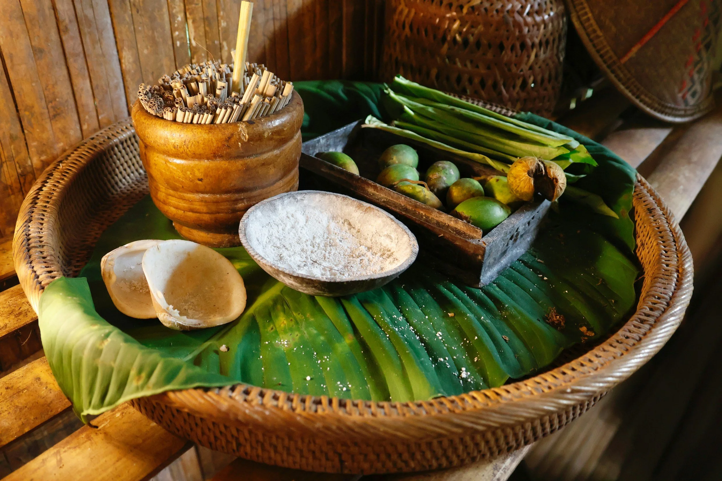 Traditional Asian ingredients on a green banana leaf, including a wooden bowl of chopsticks, a bowl of white powder, green leaves, round green fruits, and a coconut shell, set in a rustic wooden setting.