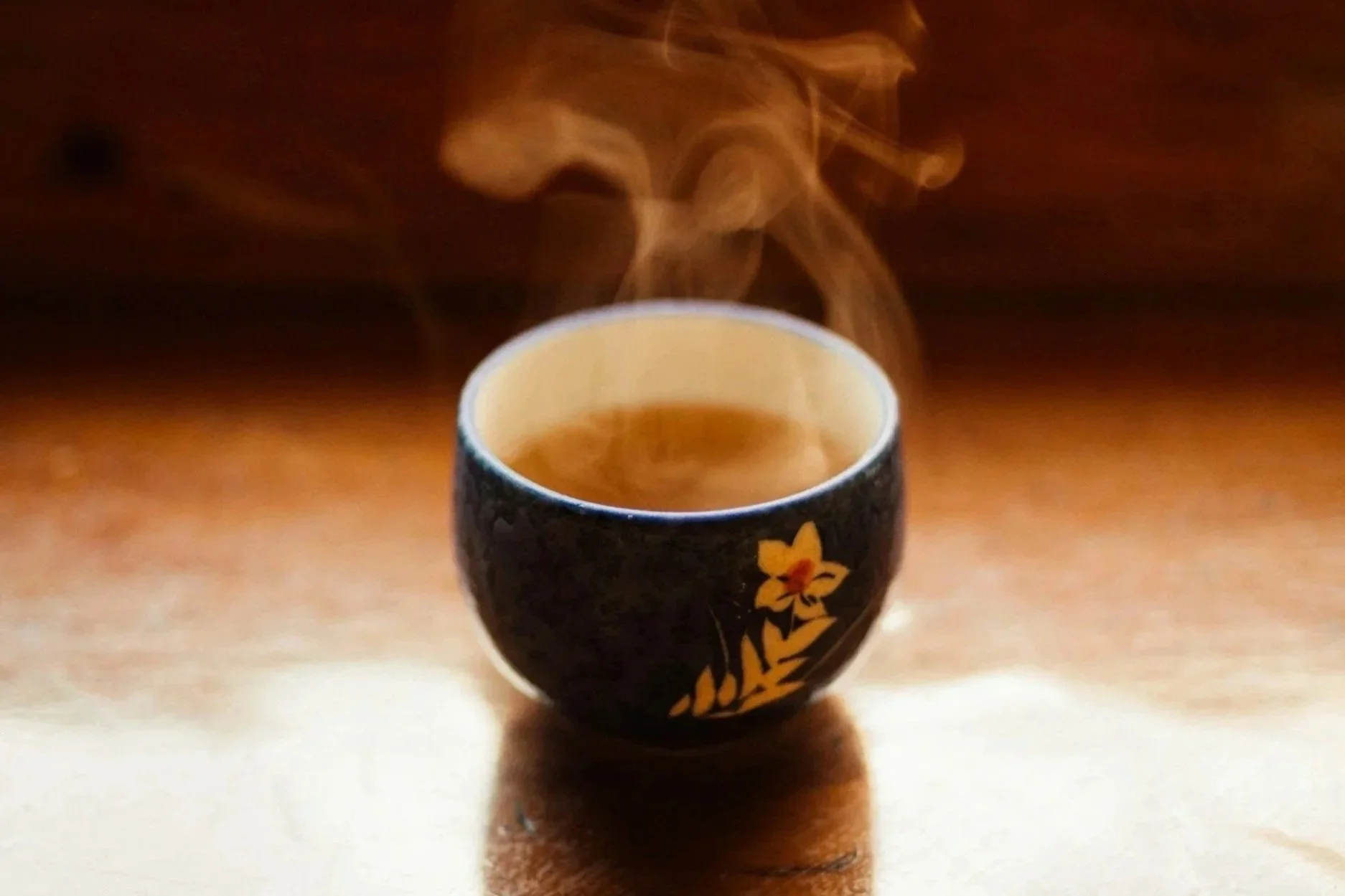 A black ceramic cup with a floral design, filled with hot tea, with steam rising from it, placed on a wooden surface.