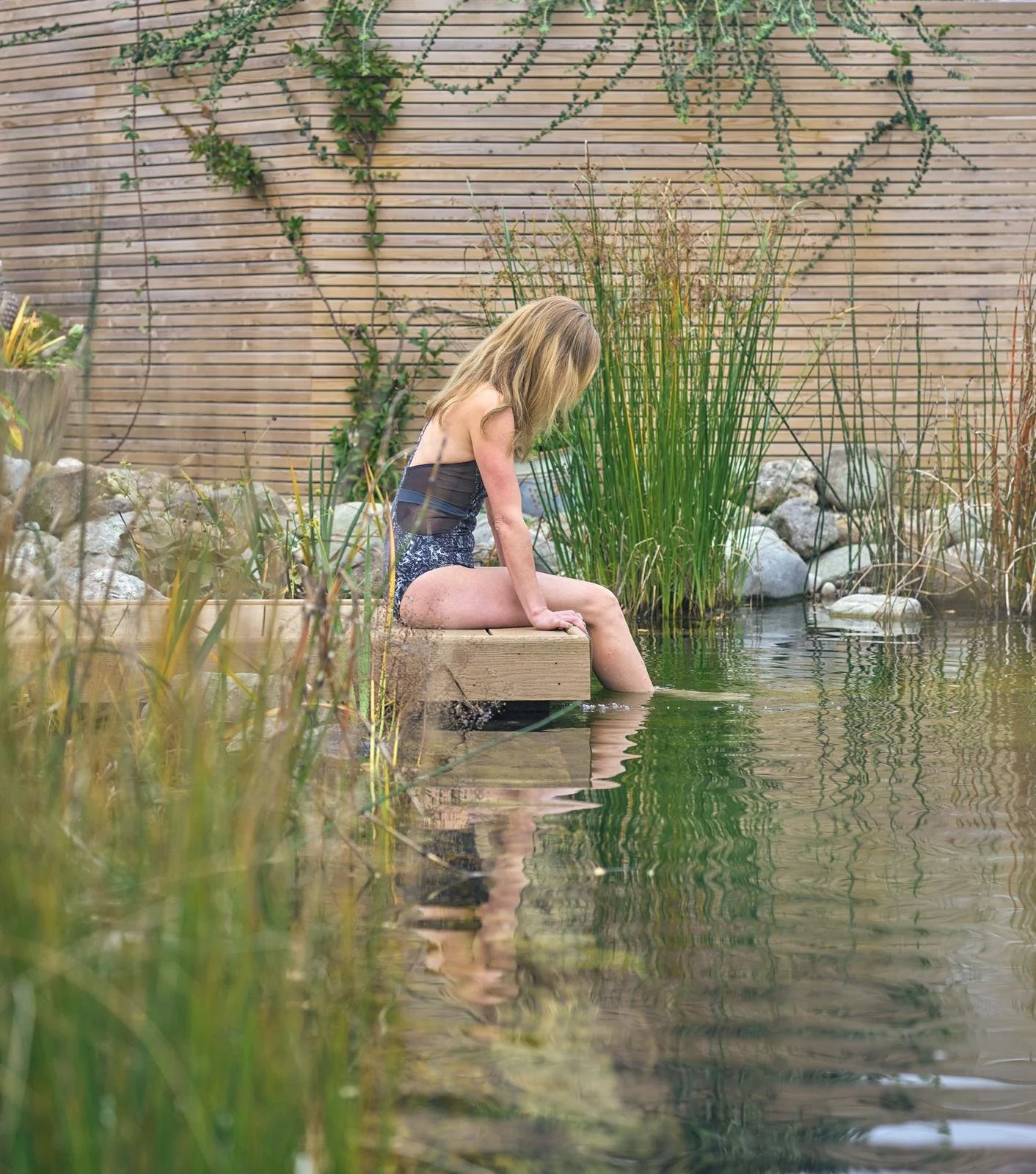 A lady sitton on a decking, with her feet in a swimming pond, wearing her swimsuit, looking calm and peaceful