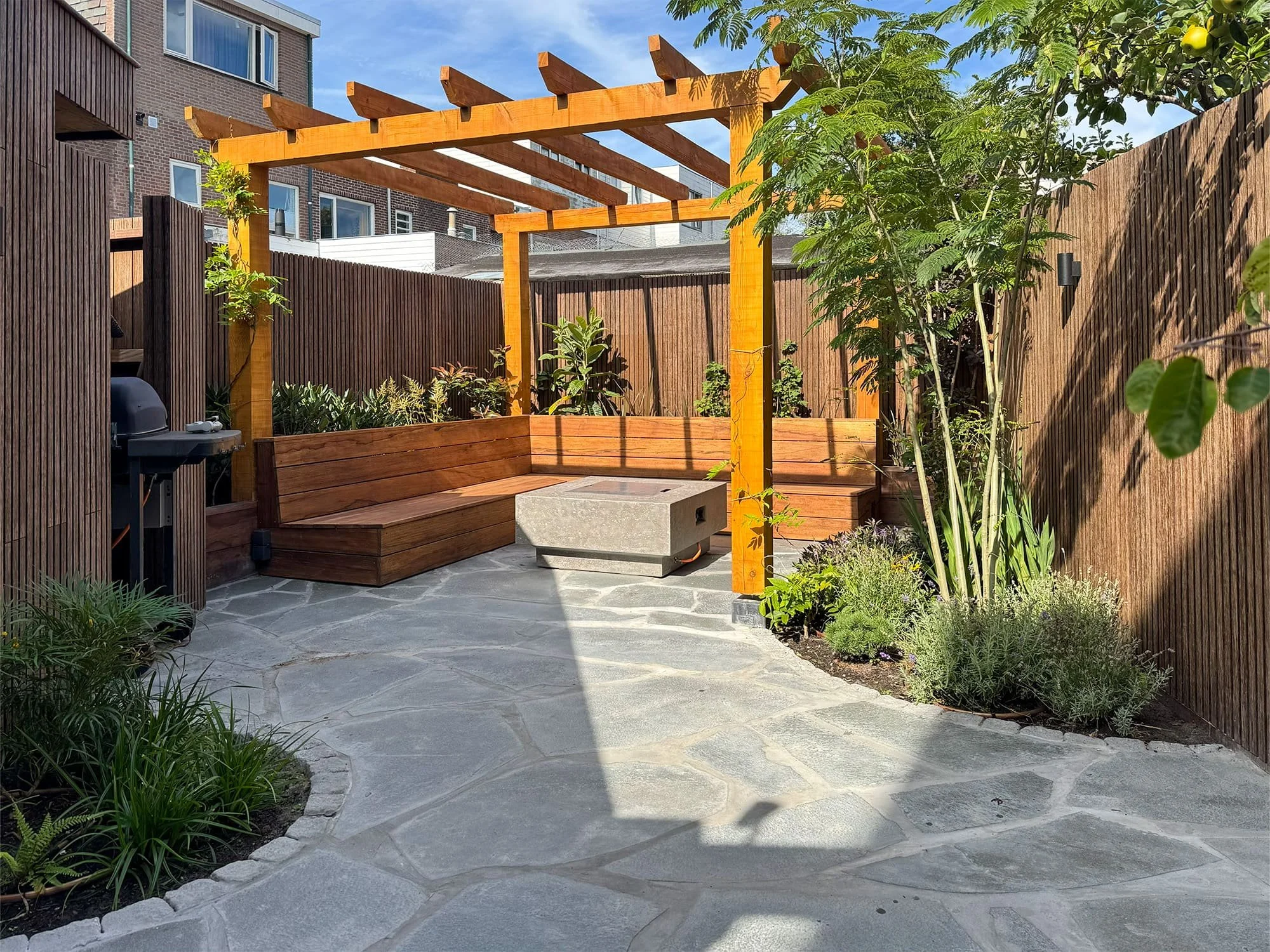 Het middelpunt van deze mediterrane achtertuin wordt gevormd door een robuuste douglashouten pergola. In combinatie met de onregelmatige flagstones als bestrating creëert dit een warme, zuidelijke sfeer die de basis vormt voor het gehele tuinontwerp.