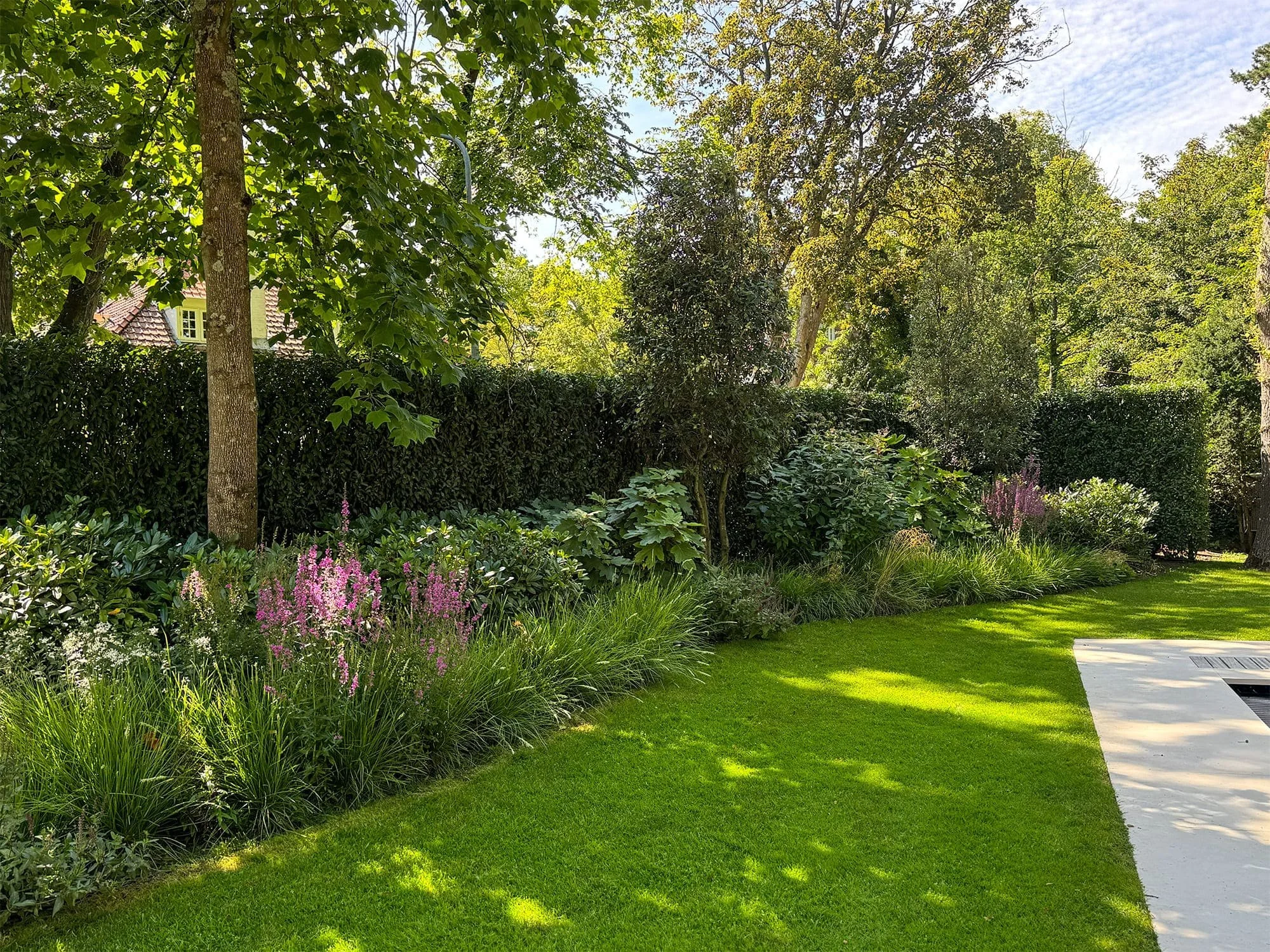 Het uitgestrekte gazon fungeert als het open, groene hart van de tuin. Door het perceel te omringen met rijkgevulde plantenborders ontstaat er een besloten sfeer die de karakteristieken van een klassieke bostuin versterkt.