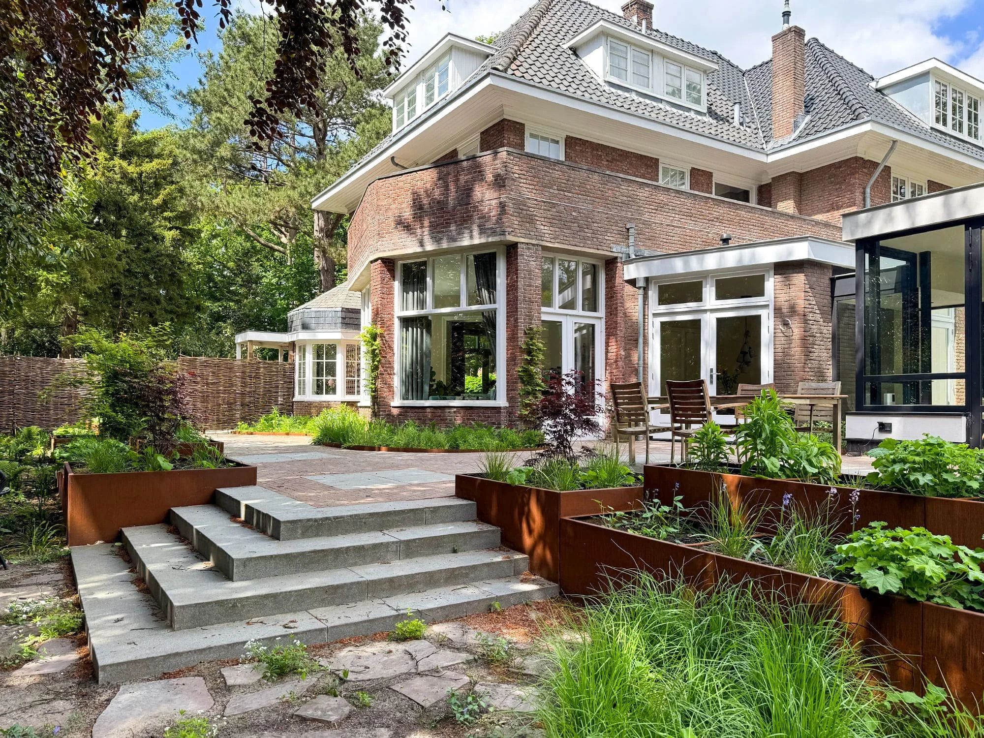 Voor deze villa heeft Studio Holtland een bostuin ontworpen. De hoogteverschillen worden opgevangen door robuuste granieten trappen en cortenstalen plantenbakken, gevuld met siergrassen zoals Sesleria. Voor de bestrating is een combinatie gebruikt va