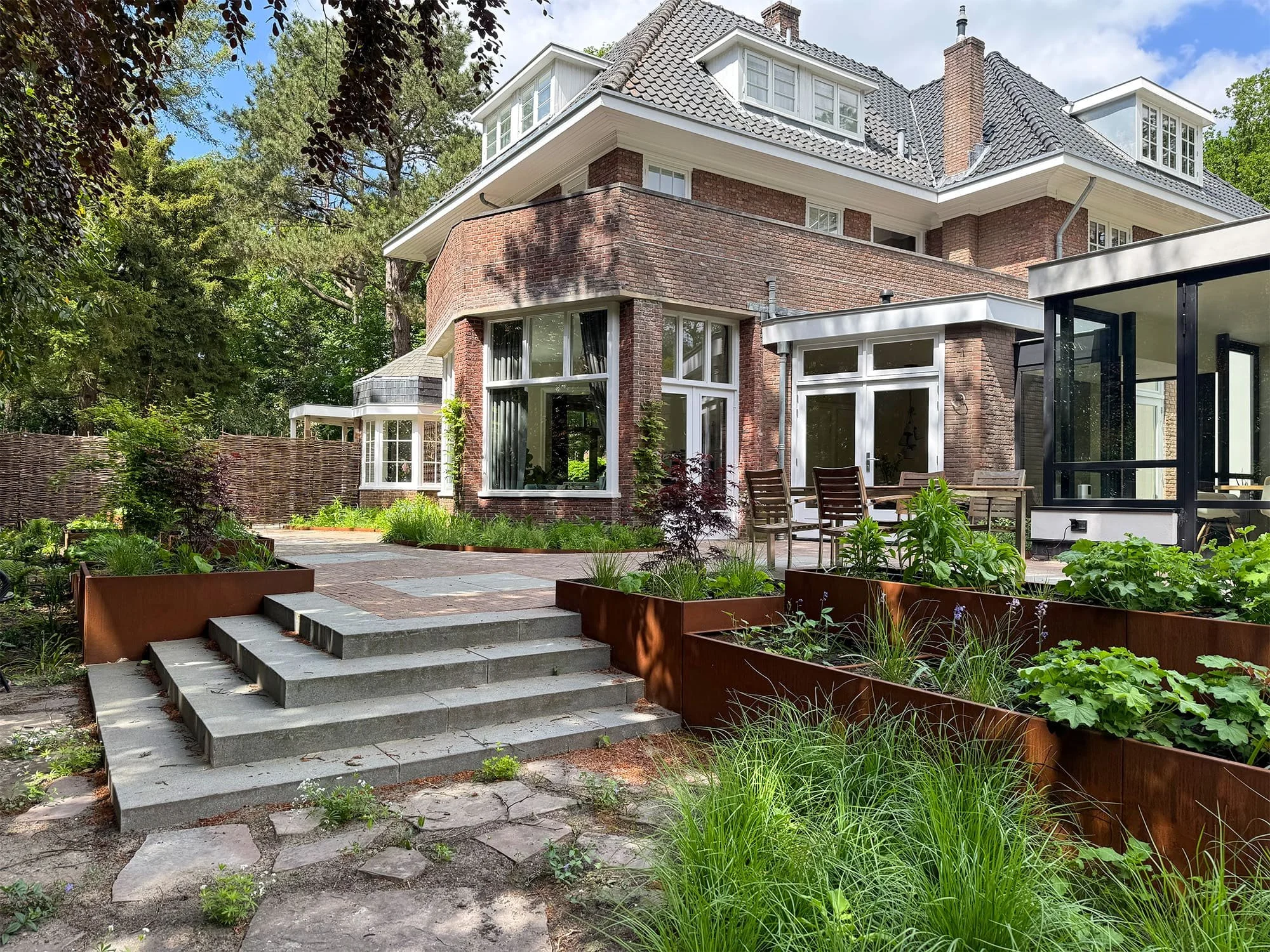 Bakstenen villa met een bostuin, voorzien van cortenstalen plantenbakken en een brede granieten trap naar het terras.