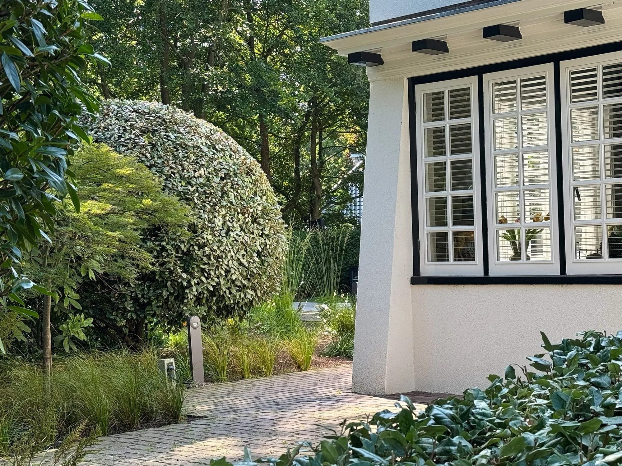 A house with white walls and large windows with white shutters, surrounded by green trees and bushes, with a stone pathway leading to the entrance.