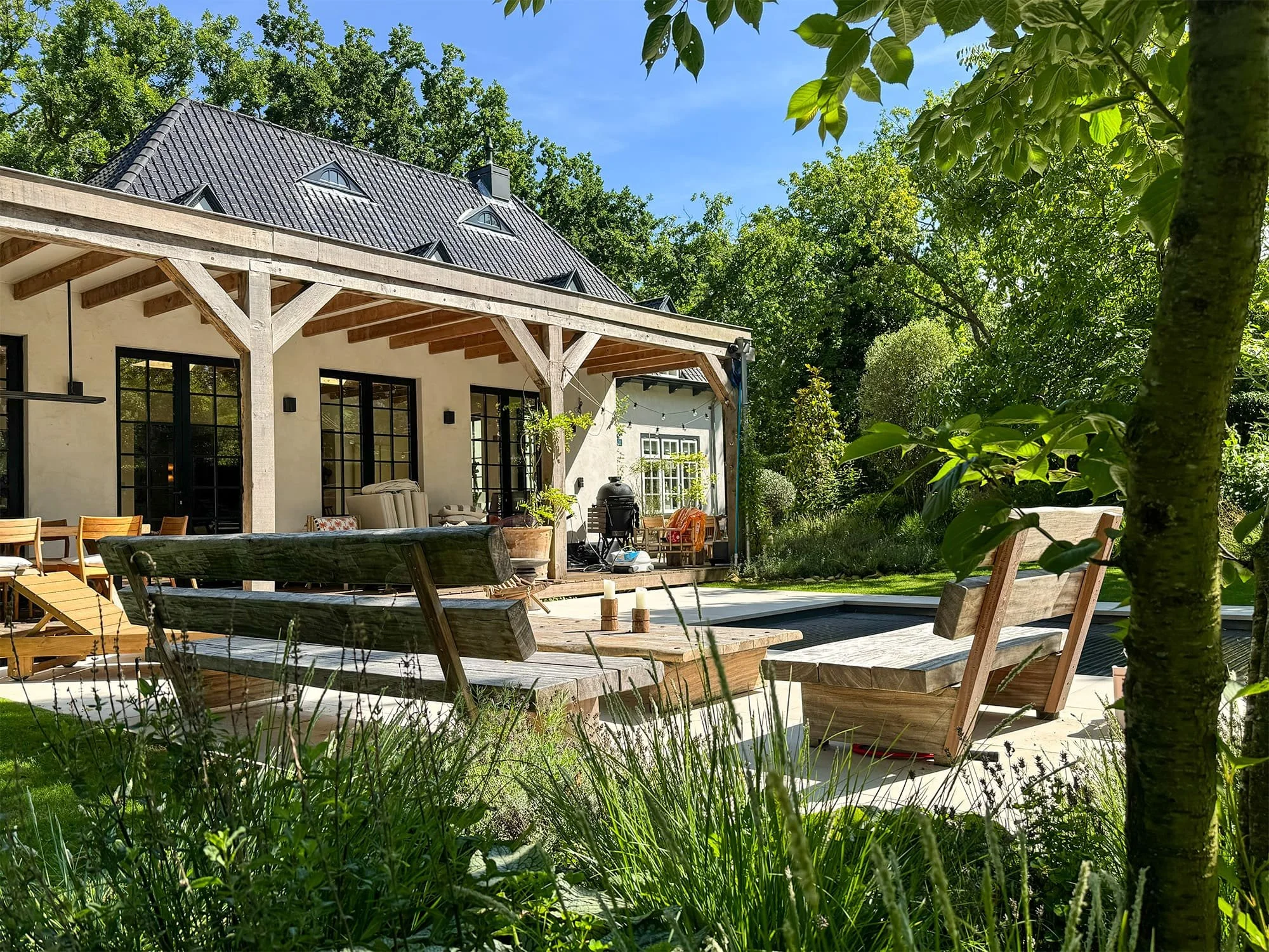 Onder de witgeschilderde veranda is een comfortabele buitenruimte gecreëerd. Vanaf het verharde terras met houten loungemeubilair is er een open en rustgevend uitzicht over het strakke gazon en het aangrenzende zwembad.