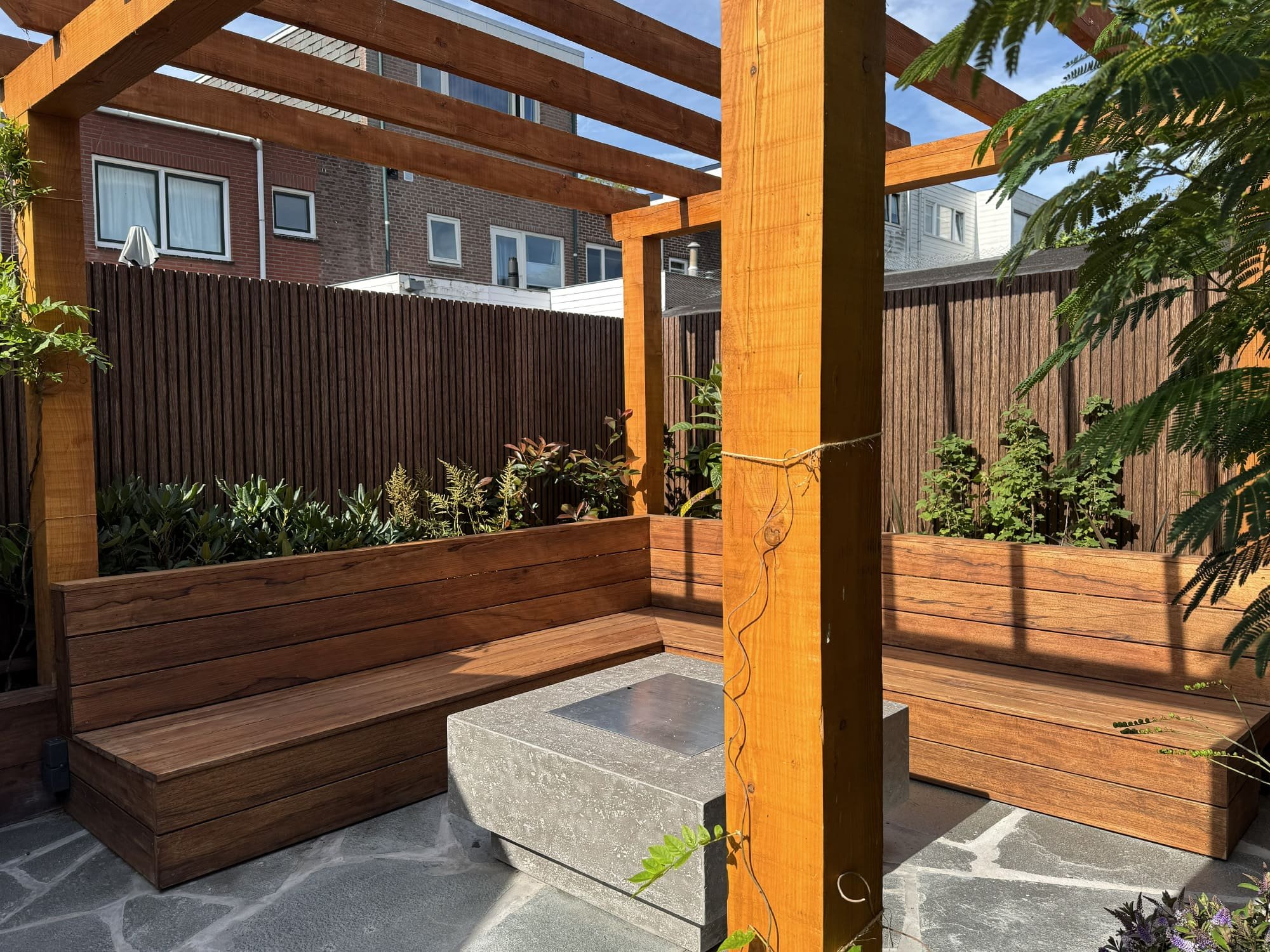 De ruime houten loungebank is naadloos geïntegreerd in de staanders van de douglashouten pergola. Deze functionele, op maat gemaakte indeling optimaliseert de beschikbare vierkante meters rondom het centrale stenen vuurelement.