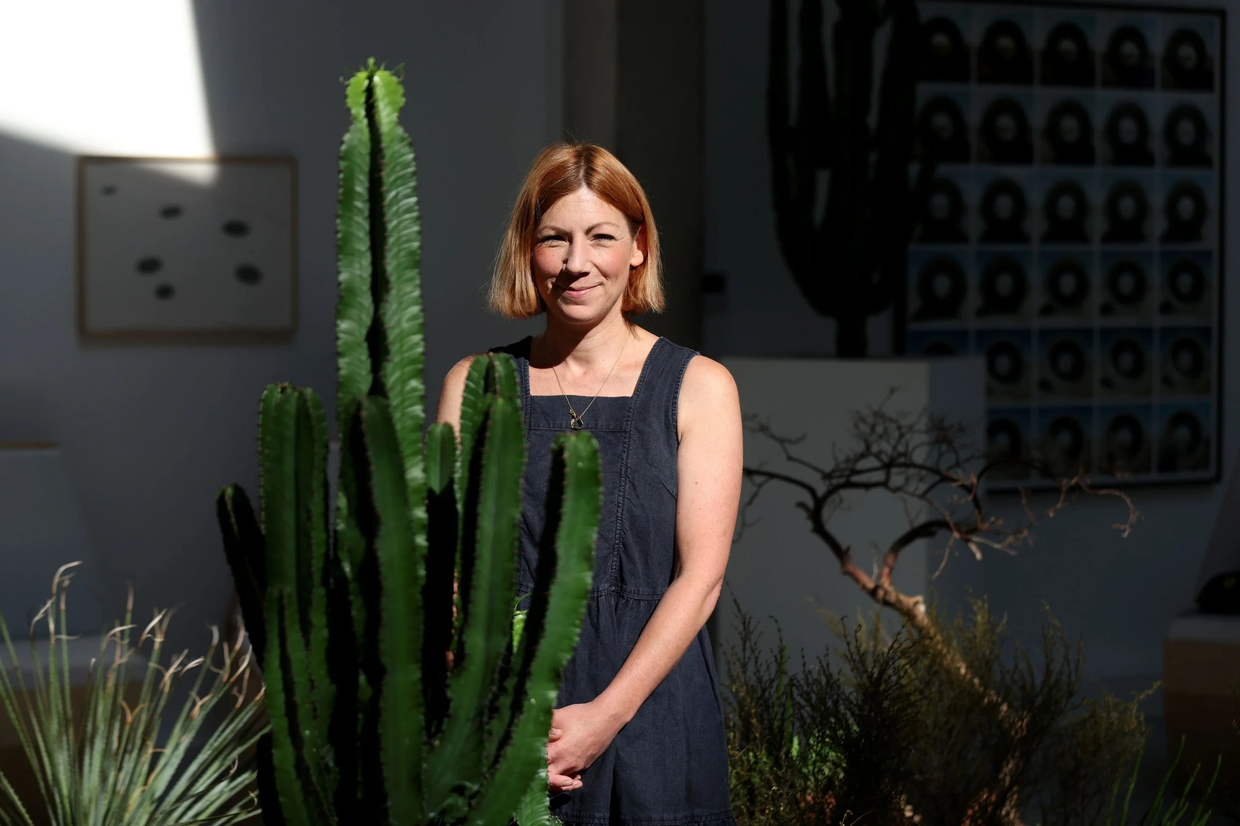 A woman with red hair standing behind various cacti, smiling in a room with artwork on the wall.