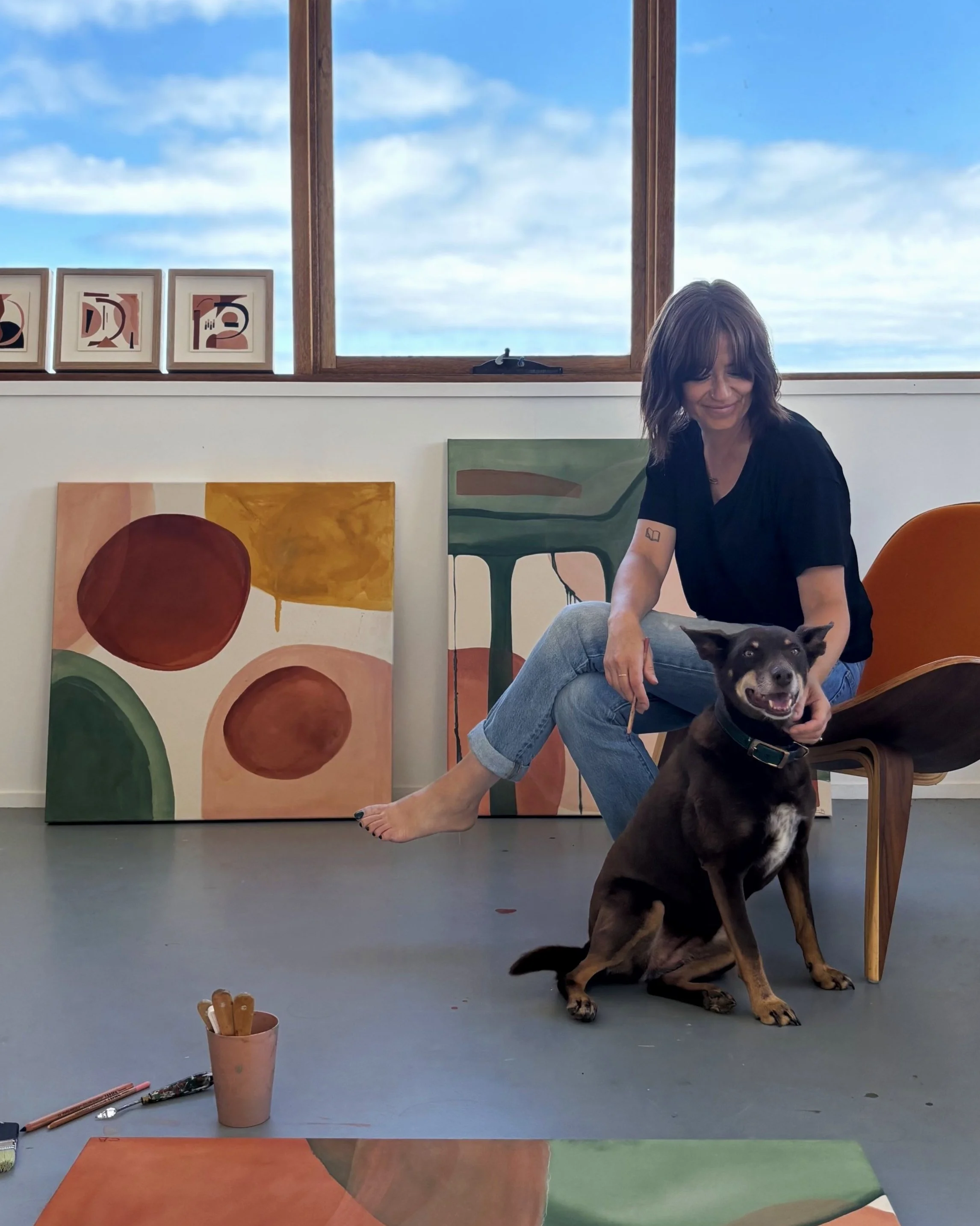Woman sitting in an art studio with abstract paintings, accompanied by a dog. She is barefoot and wearing a black shirt and jeans. The room has a large window showing a blue sky with clouds.