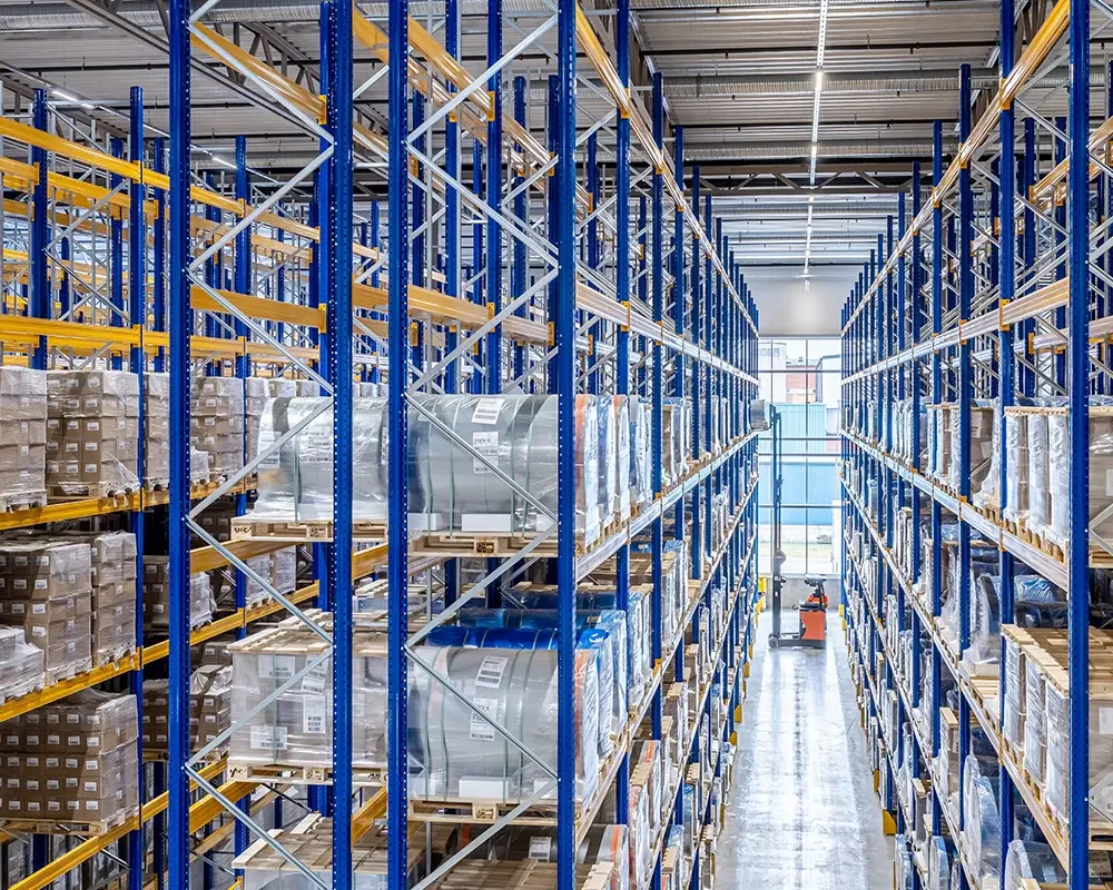 View of a warehouse aisle with tall blue and yellow shelving filled with boxed and wrapped products.