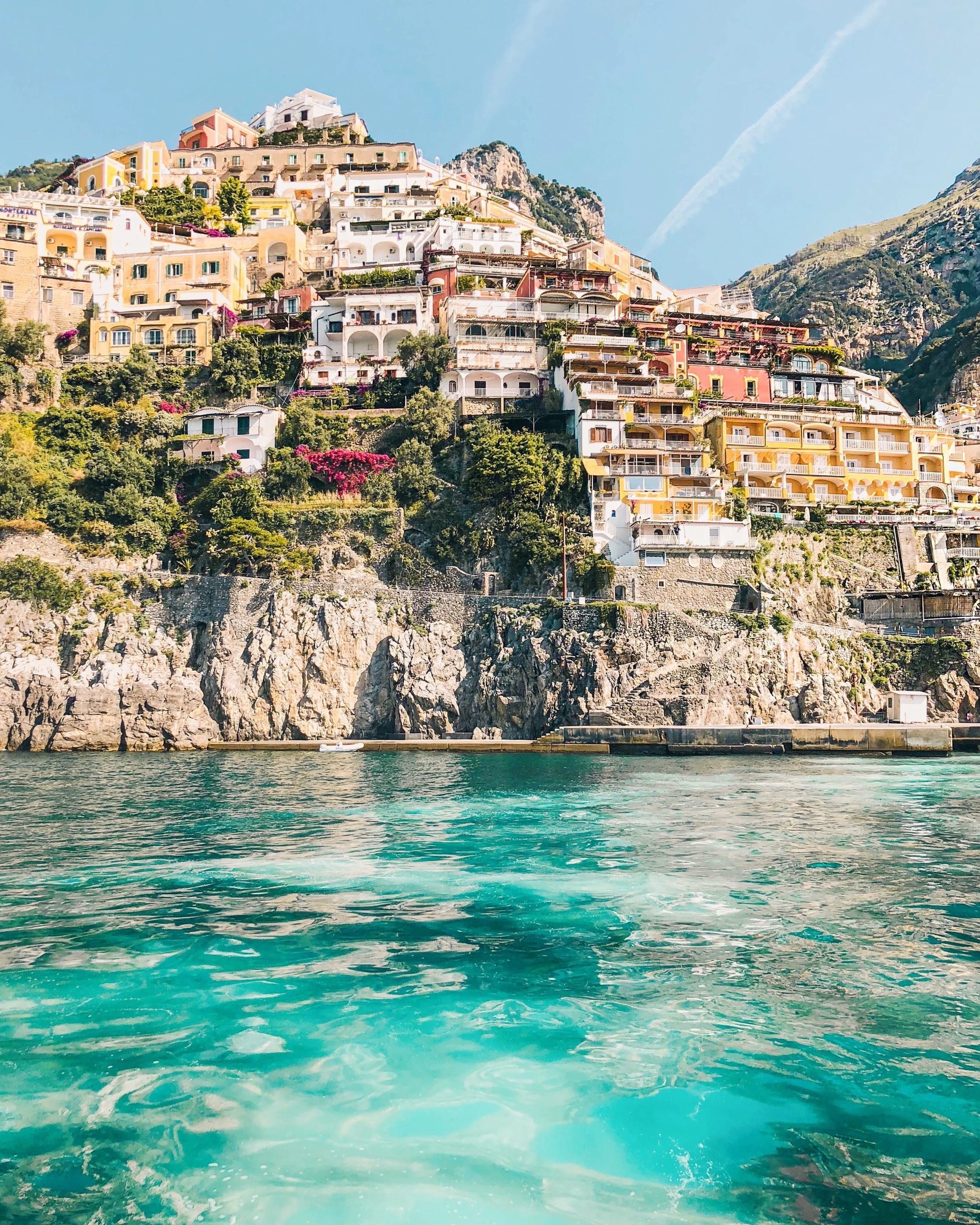 Colorful buildings and houses built into a steep hillside on a coastal cliff, overlooking turquoise waters with a small boat, in Positano, Italy.