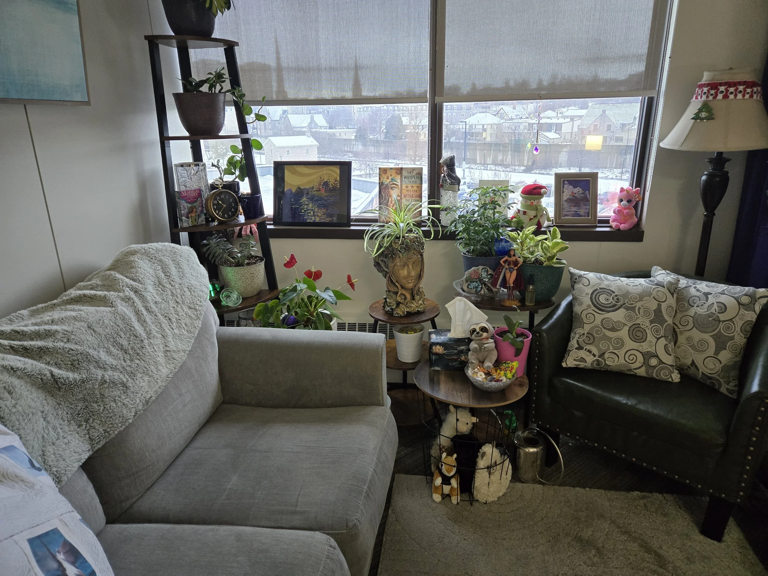 Living room with a gray sofa, black armchair with patterned pillows, wooden side tables filled with plants, decorative items, and plush toys, large window with a snow-covered outdoor scene, and a tall black floor lamp.