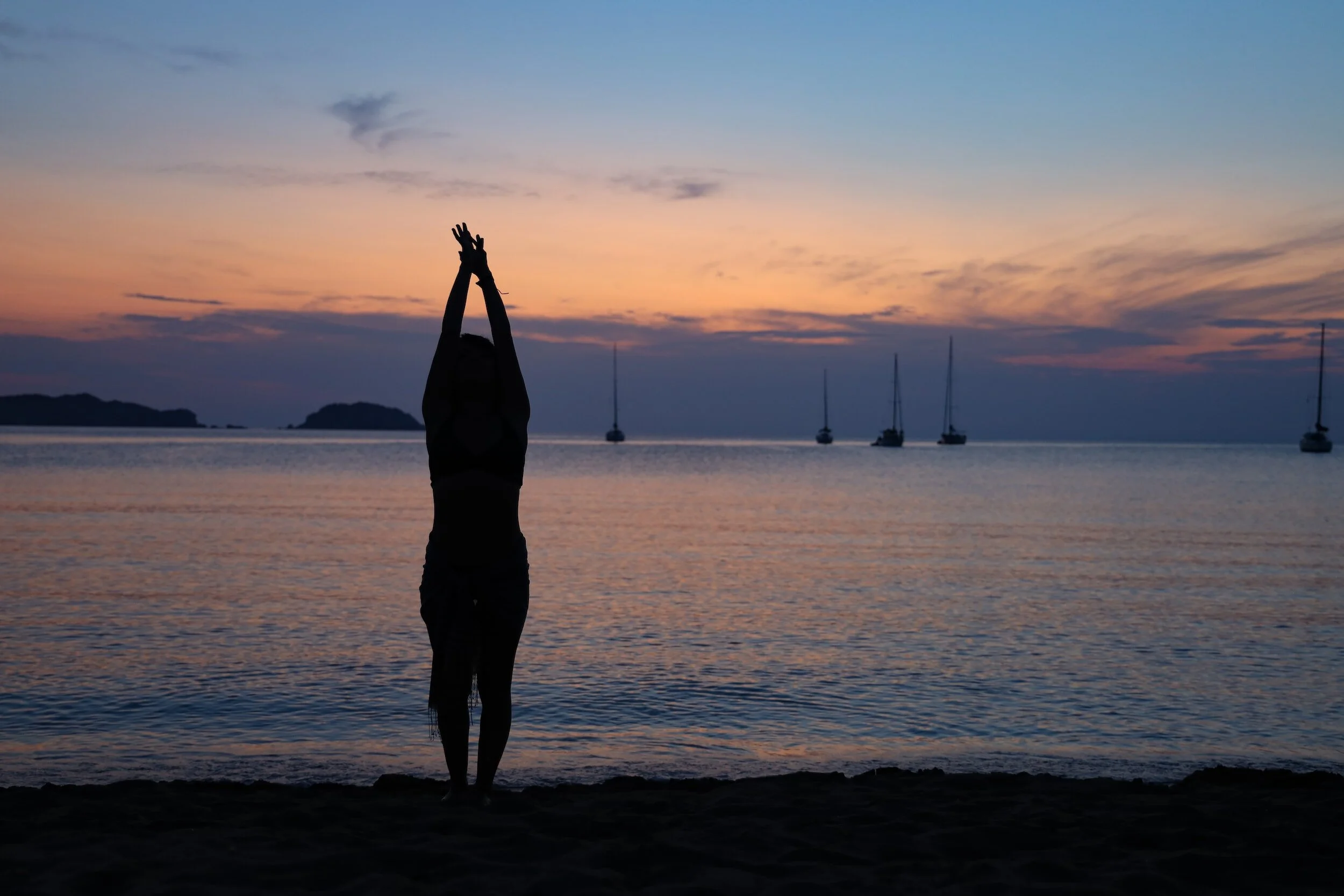 Silhouette of a person stretching on a beach at sunset with sailboats on the horizon.