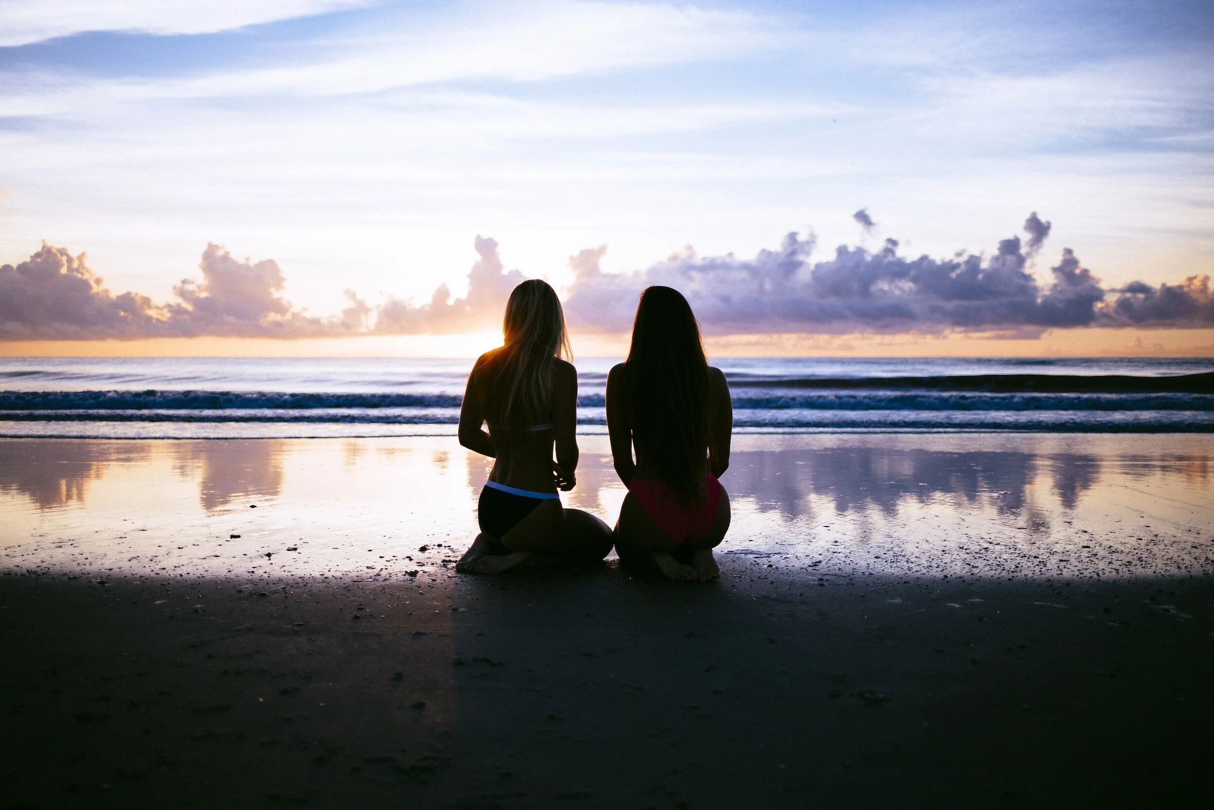 Two women kneeling on a beach watching the sunset over the ocean.