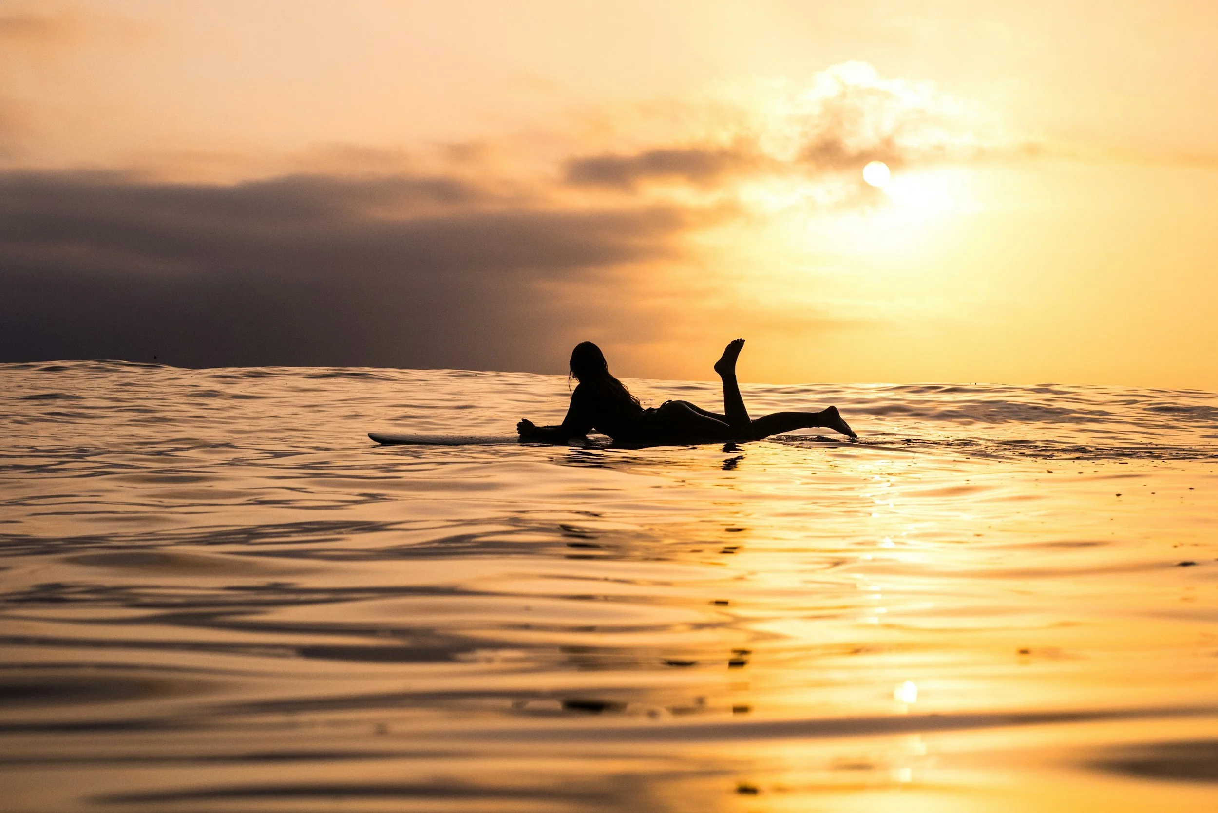 Silhouette of a person lying on a surfboard on the ocean at sunset, with a golden sky and calm water.