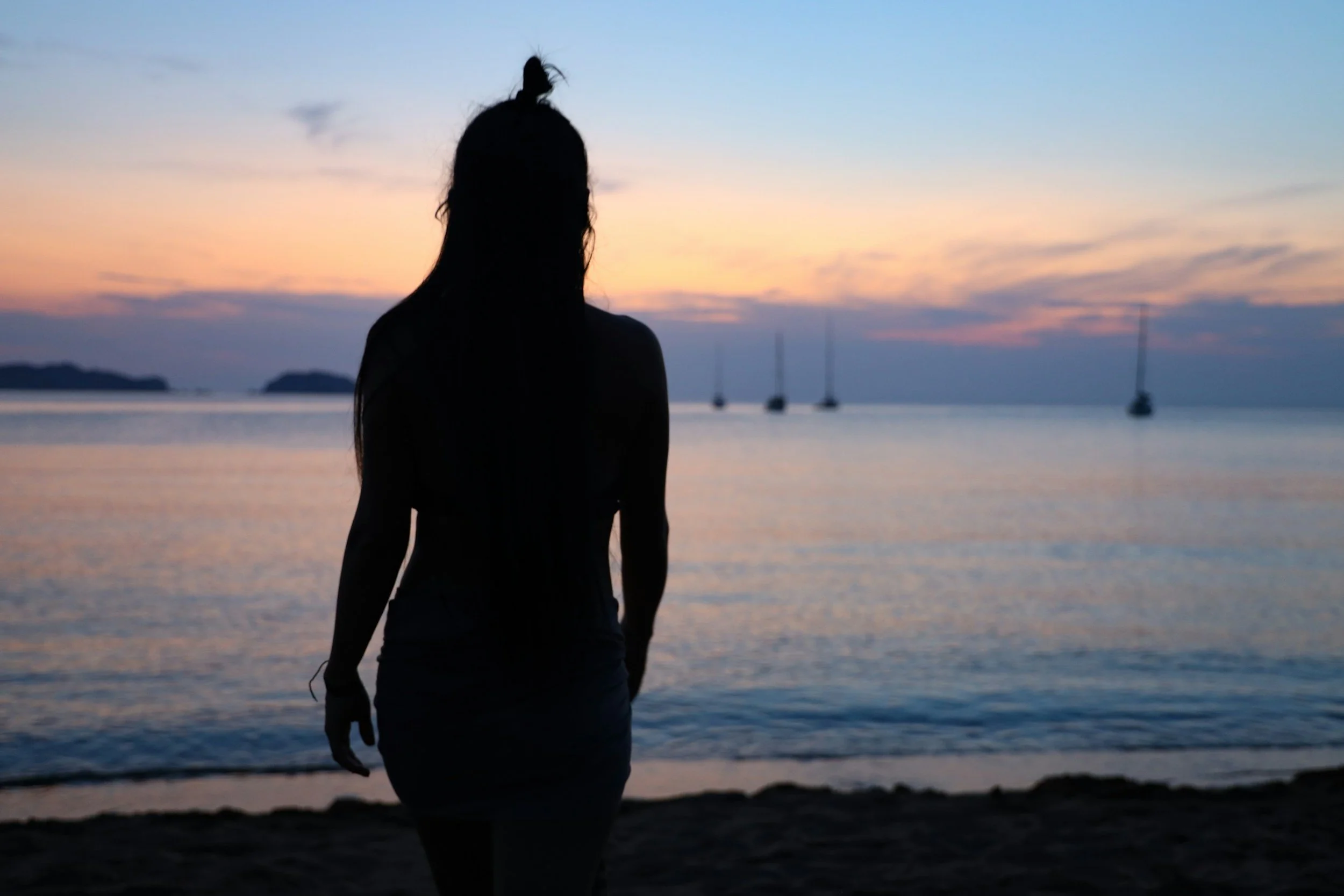 Silhouette of a woman on a beach at sunset with sailboats in the distance.