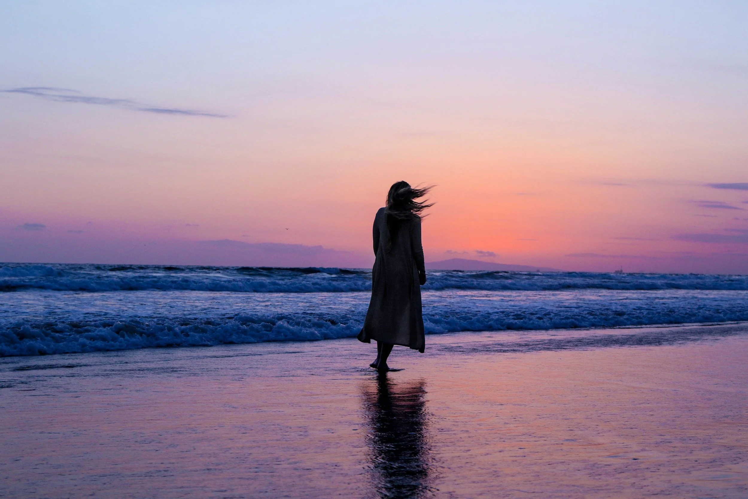 Silhouette of a person walking on the beach during a colorful sunset with waves in the background.