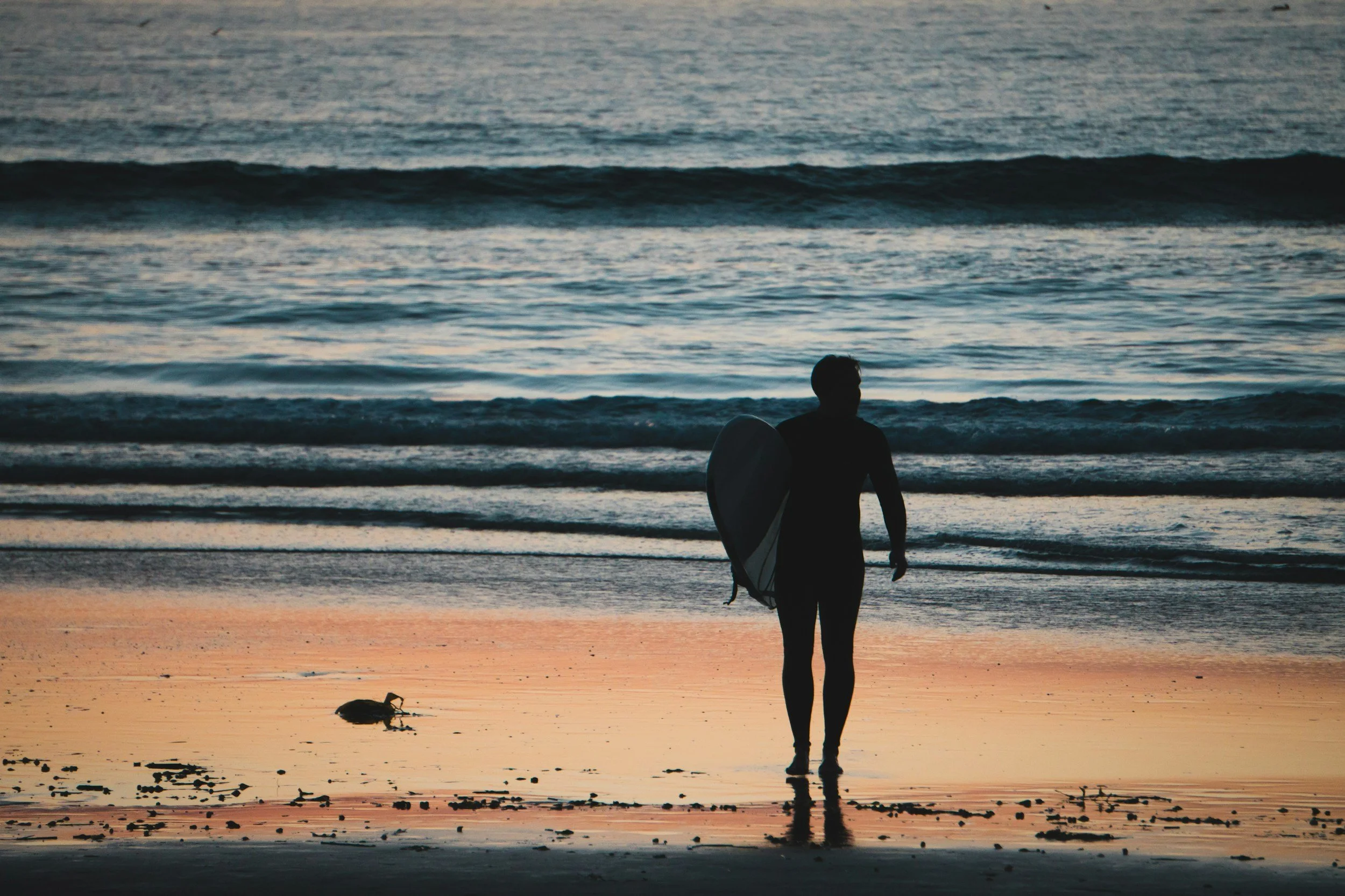 Silhouette of a person holding a surfboard on a beach at sunset with waves in the background.