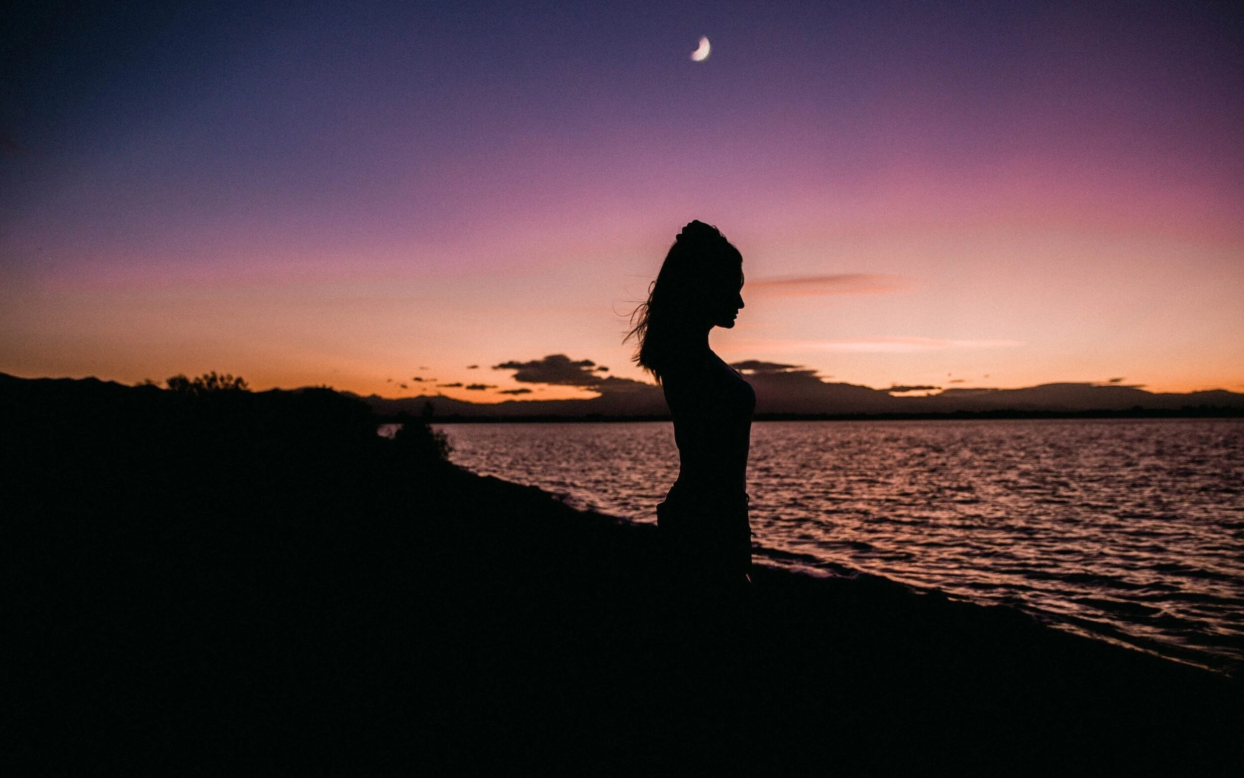 Silhouette of a woman standing by a lake at sunset with a crescent moon in the sky.