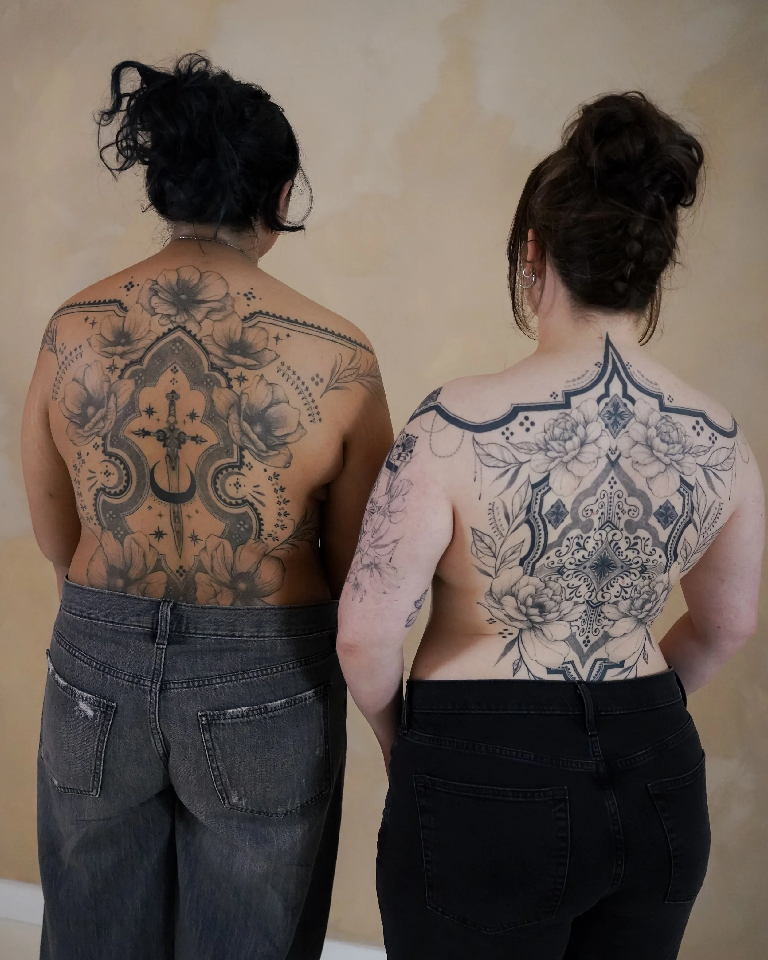 Two women with large floral tattoos on their backs, standing with their backs facing the camera, against a beige wall.