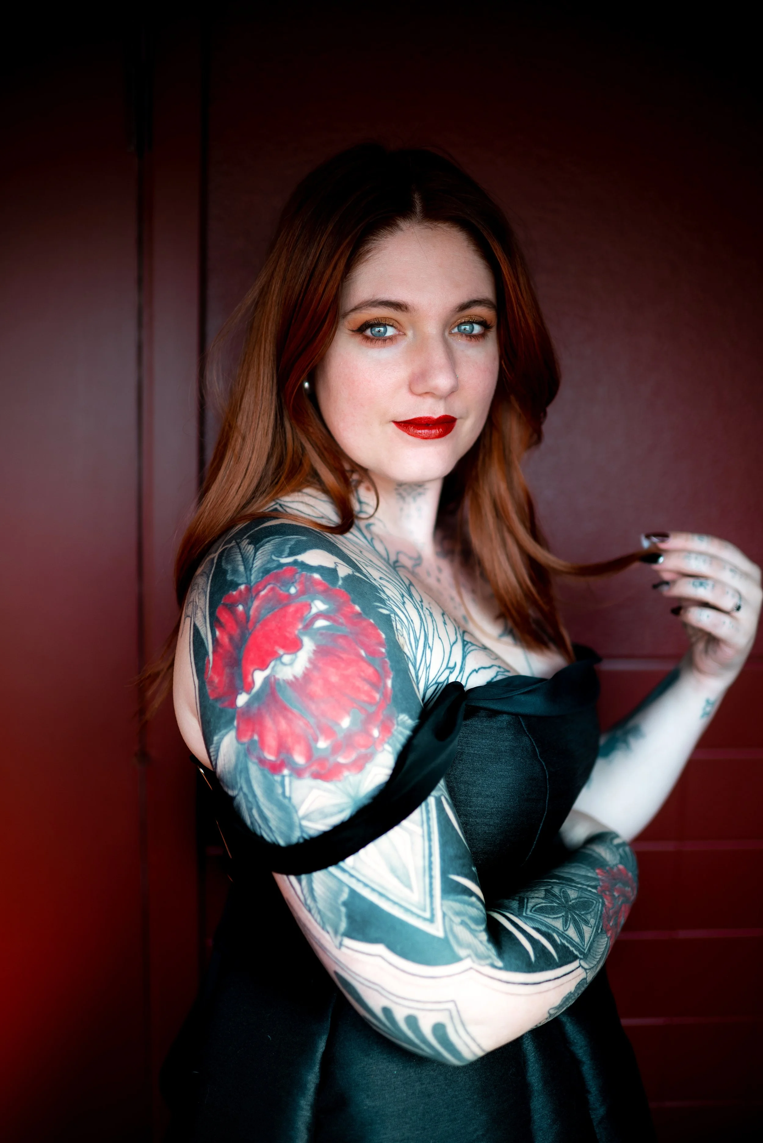 A young woman with red hair, blue eyes, and red lipstick, wearing a black off-the-shoulder dress, models with visible tattoos on her arm and chest, standing against a dark red background.