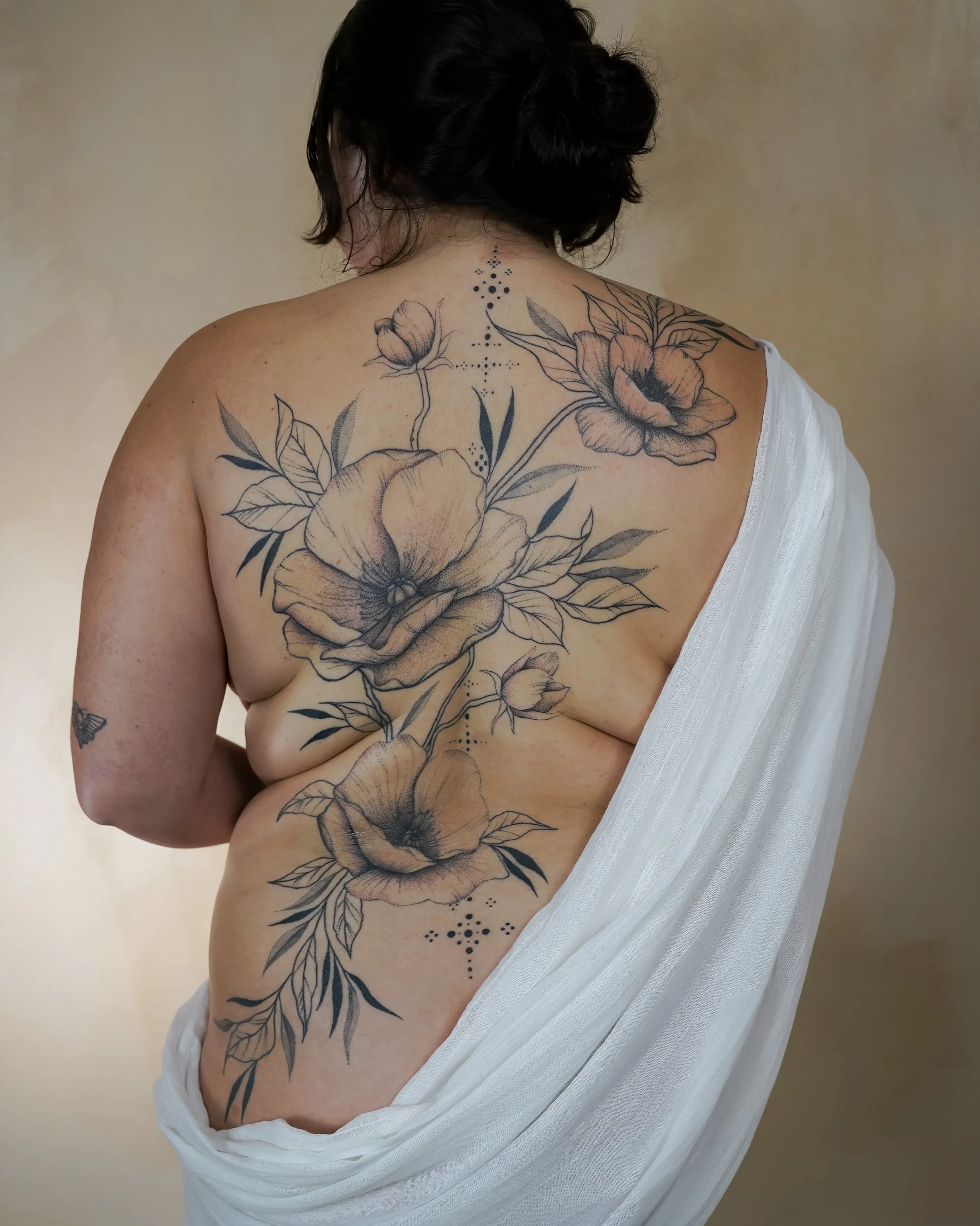 A woman with dark hair in an updo, partially covered by a draped white fabric, showcasing a large floral tattoo on her back and side, featuring detailed black and gray flowers and leaves.