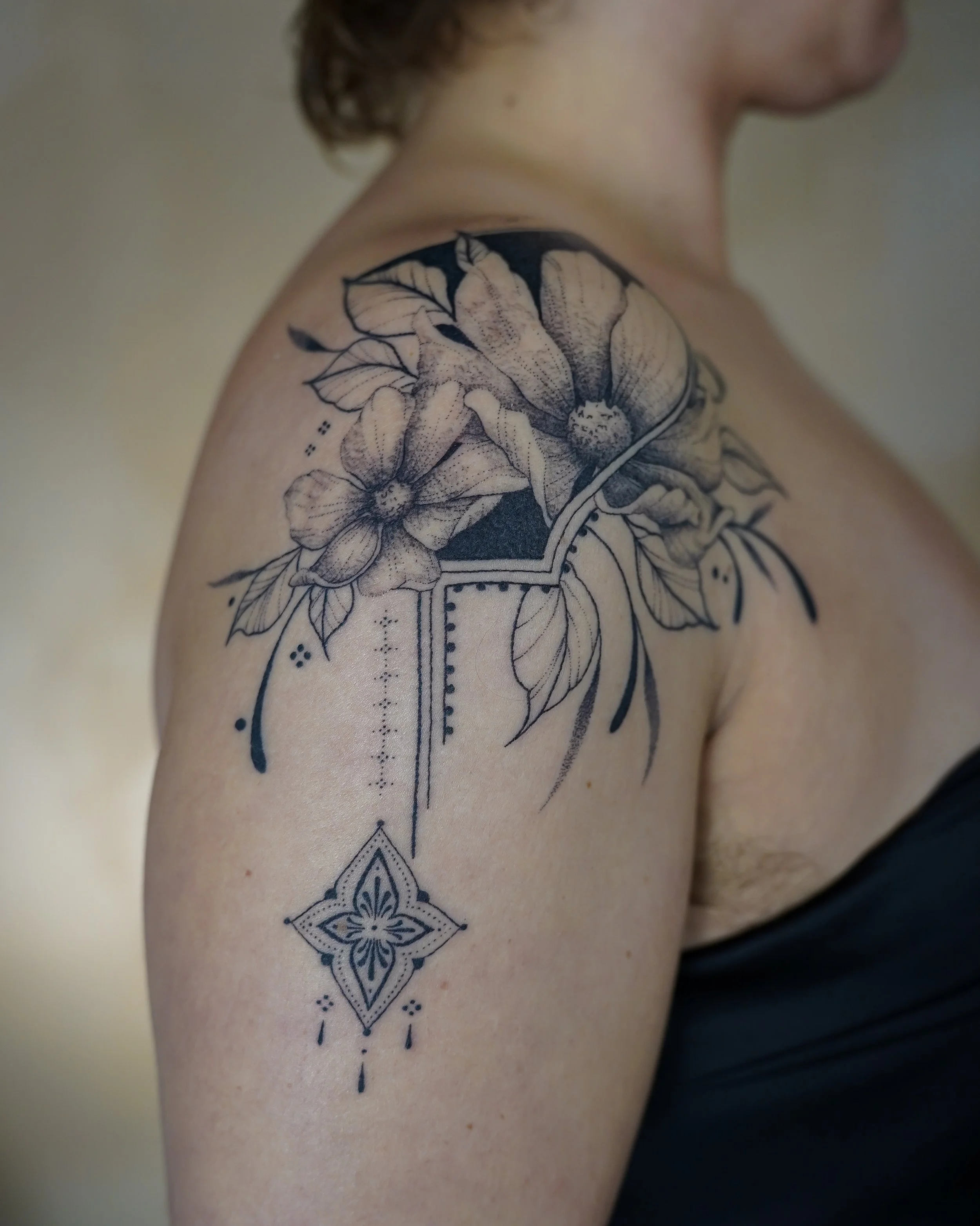 Black and gray floral tattoo featuring large flowers and leaves on the person's upper arm with decorative geometric elements hanging down.