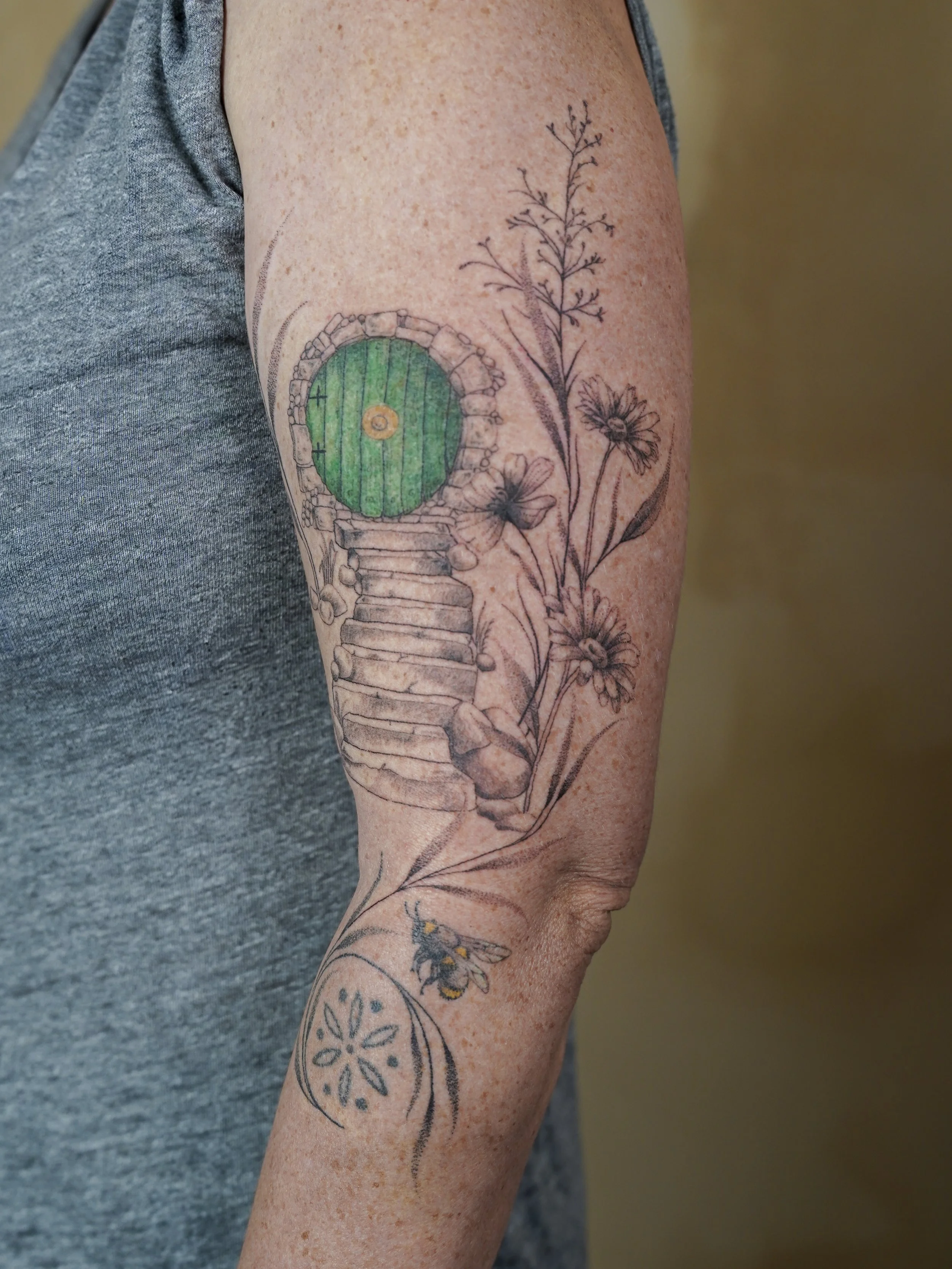 Tattoo of a Hobbit-style door and staircase with flowers and a bee on the arm.