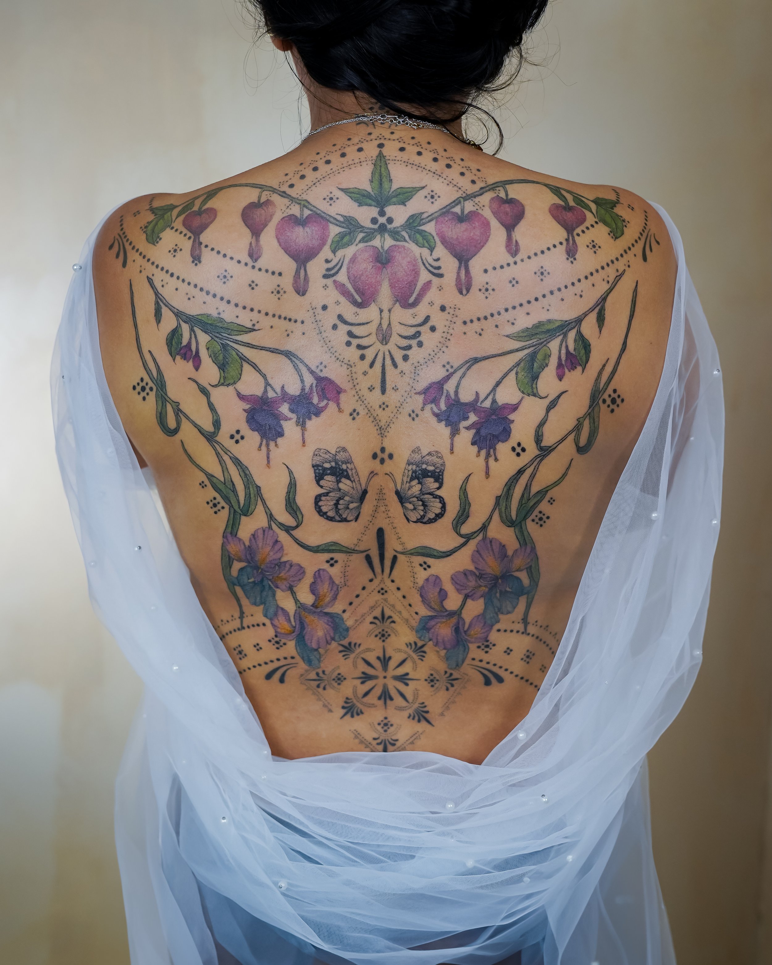 Back view of a person with a large floral and butterfly tattoo covering their back, partially draped in sheer white fabric.