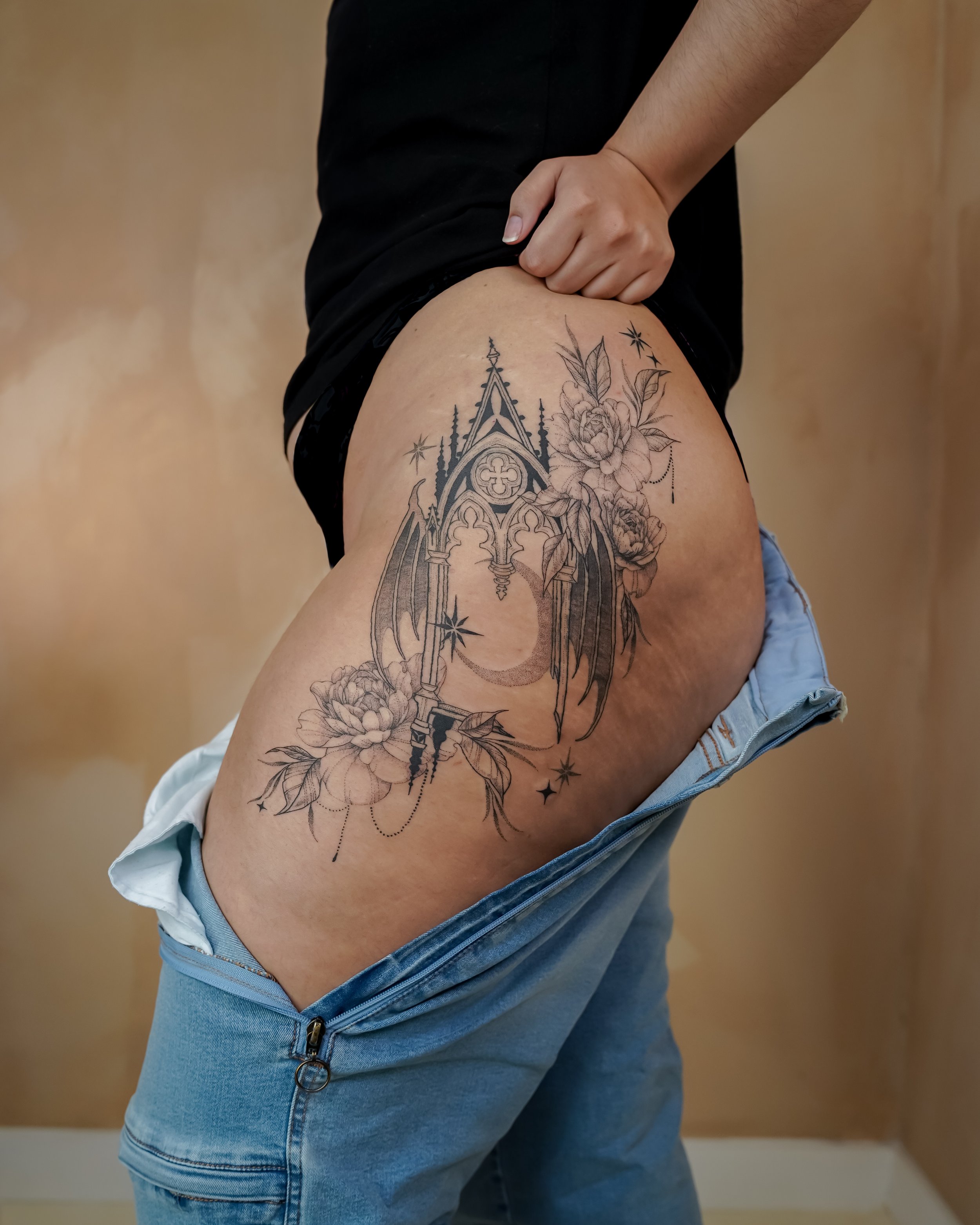 Person lifting their shirt to reveal a tattoo of a gothic cathedral with floral and celestial elements on their thigh.