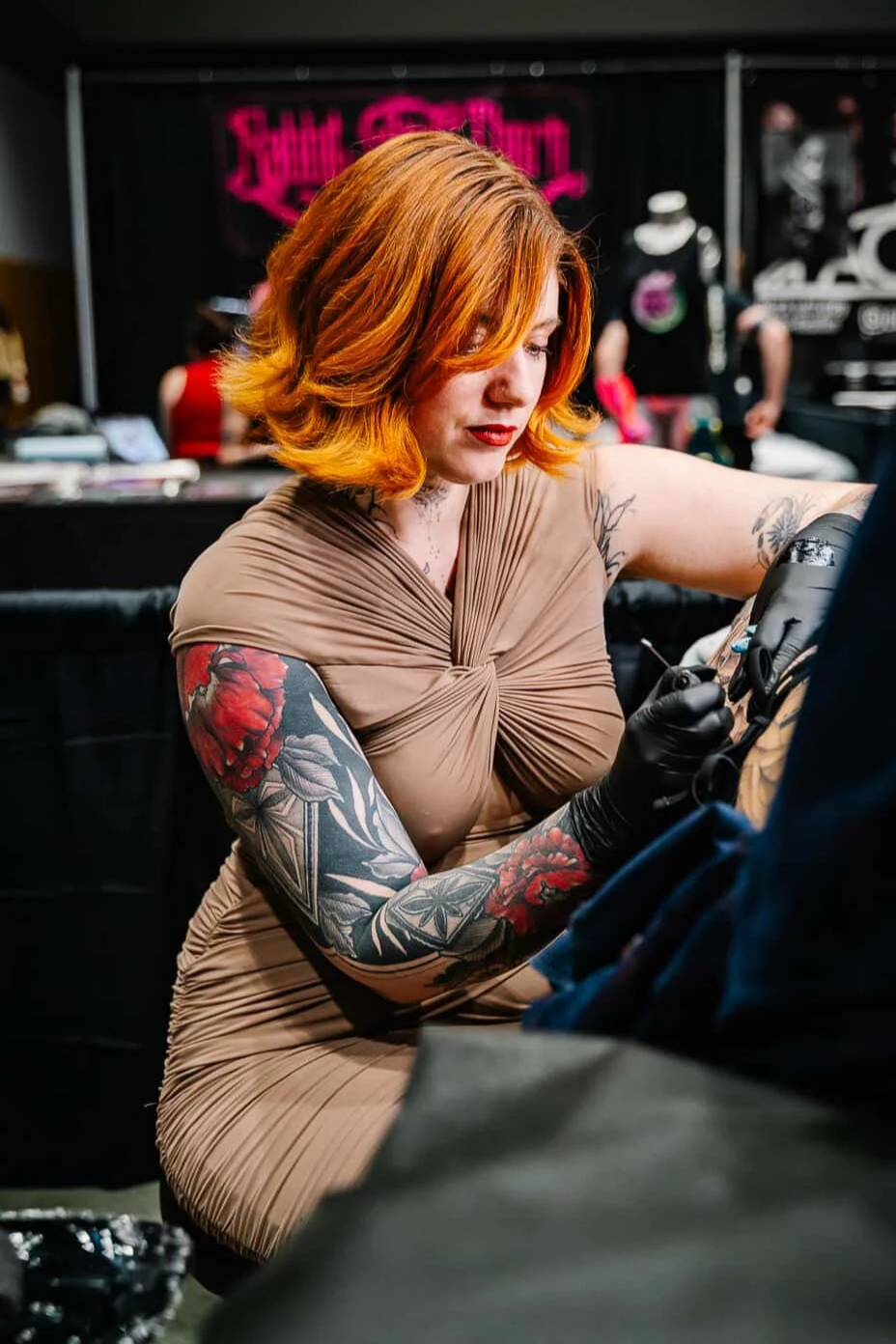 A woman with red-orange hair and tattoos on her arms tattooing.