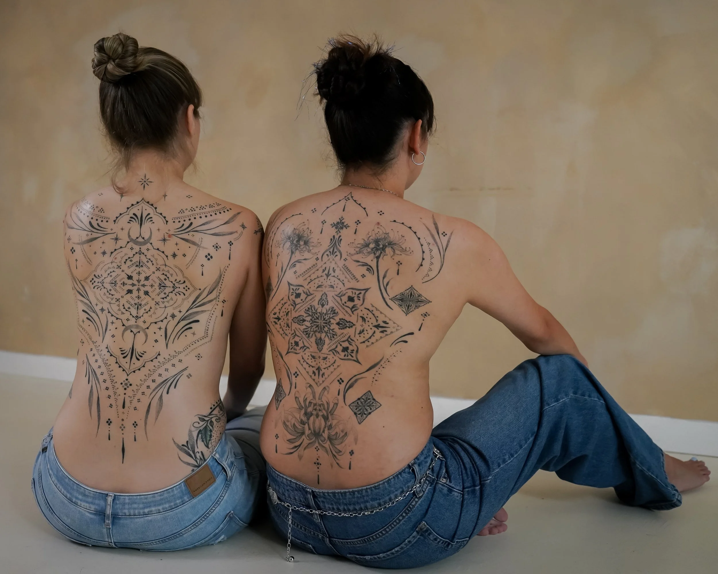 Two women with large intricate black tattoos on their backs sitting on the floor with their backs to the camera.