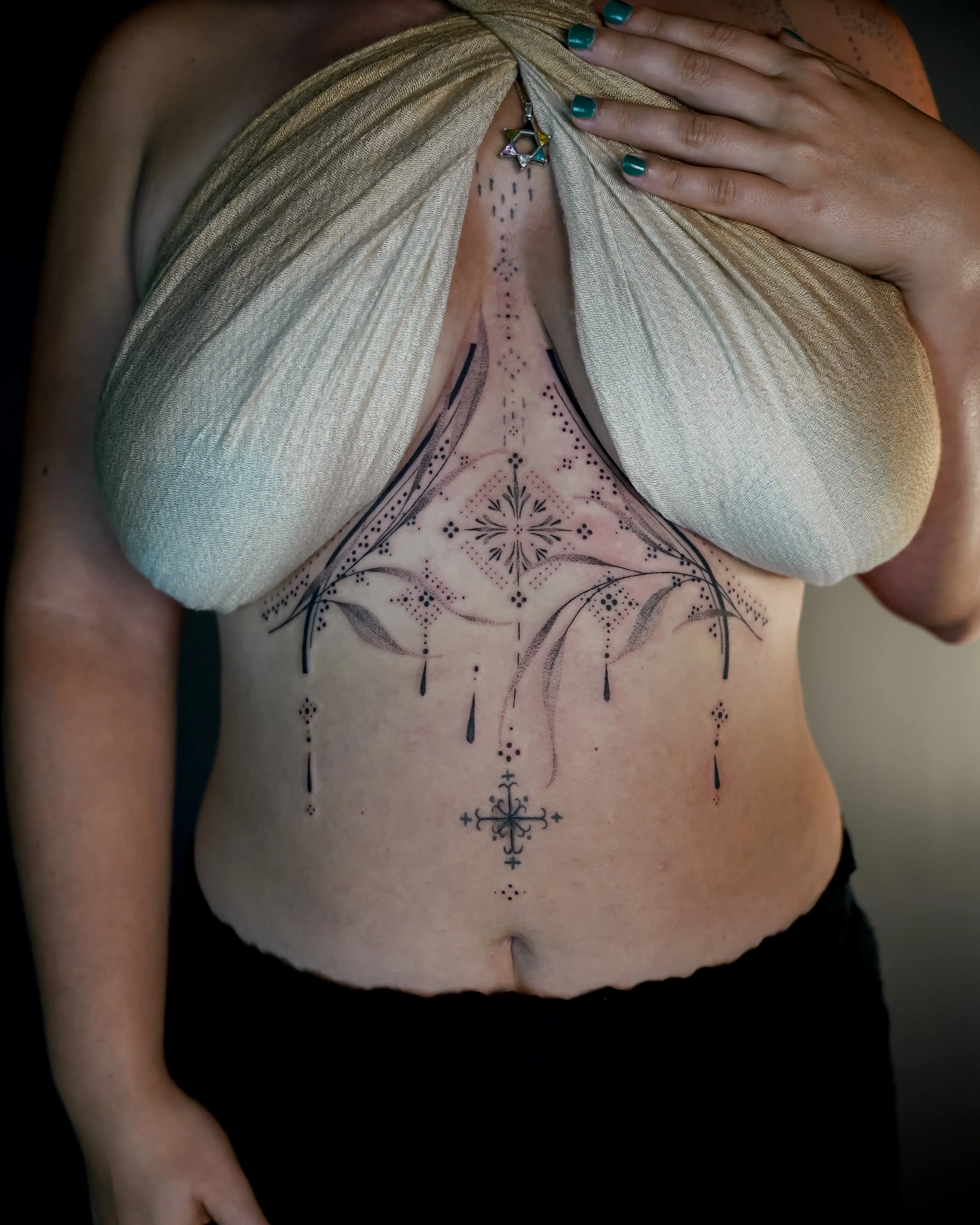 Woman with a geometric tattoo on her chest and abdomen, partially covered by a gold top and her hand with teal nail polish.