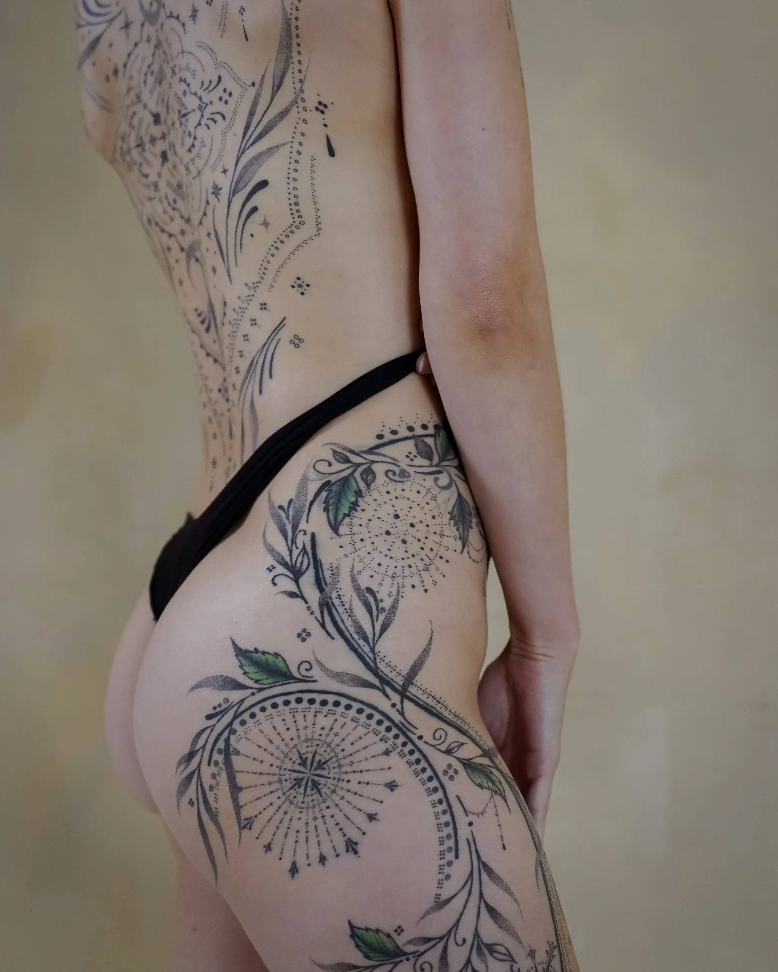 A person with elaborate black and green tattoos on their side, thigh, and arm, wearing black thong underwear, standing against a light yellow background.