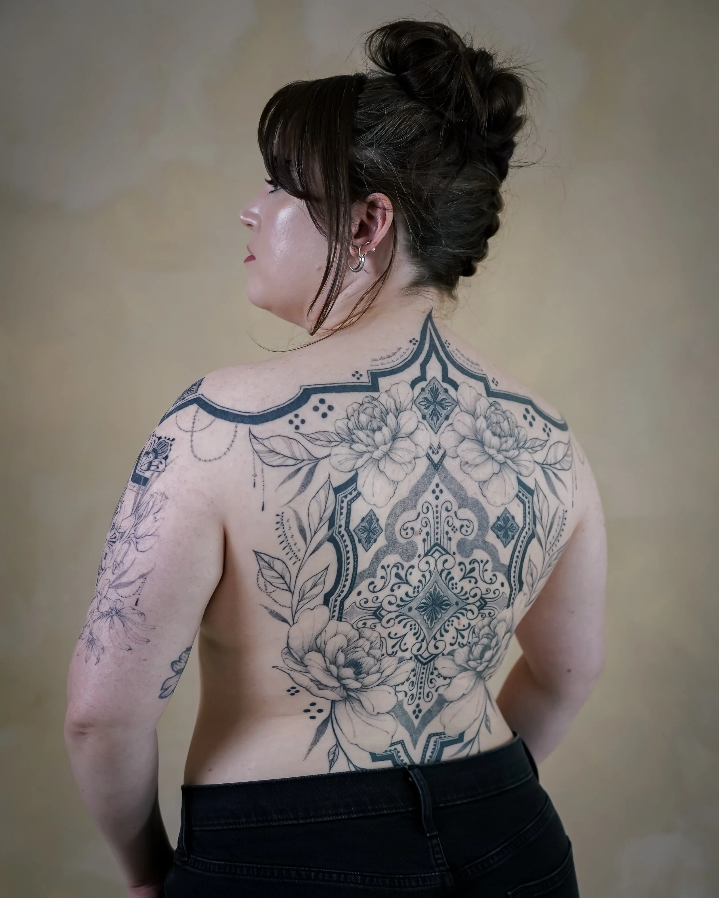A woman with dark hair in an updo, showing tattoos of flowers and geometric patterns on her back and arms, wearing earrings and black pants.