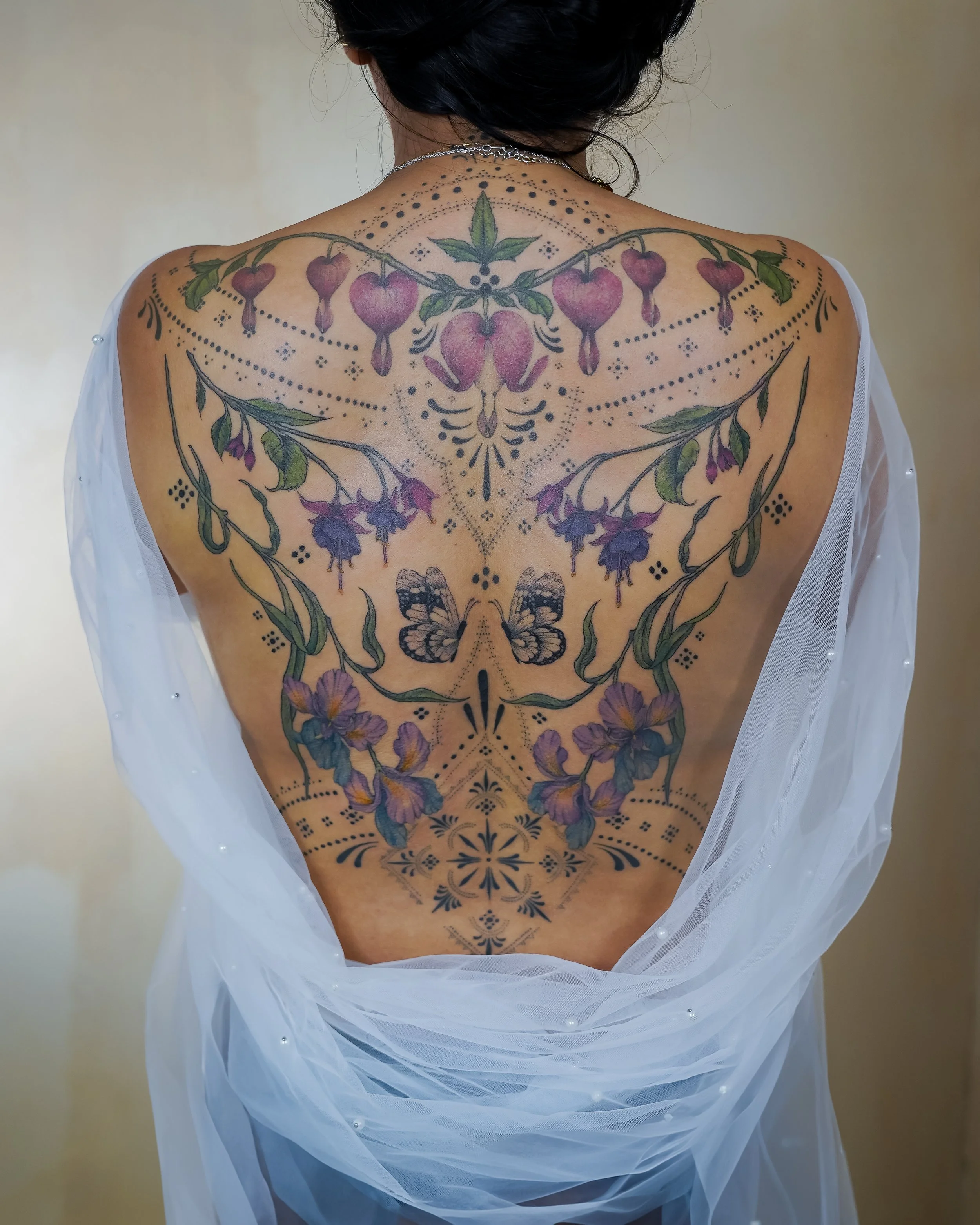 Back view of a person with a large, intricate floral tattoo covering the entire back, featuring flowers, butterflies, and decorative patterns, partially covered by a sheer white fabric.