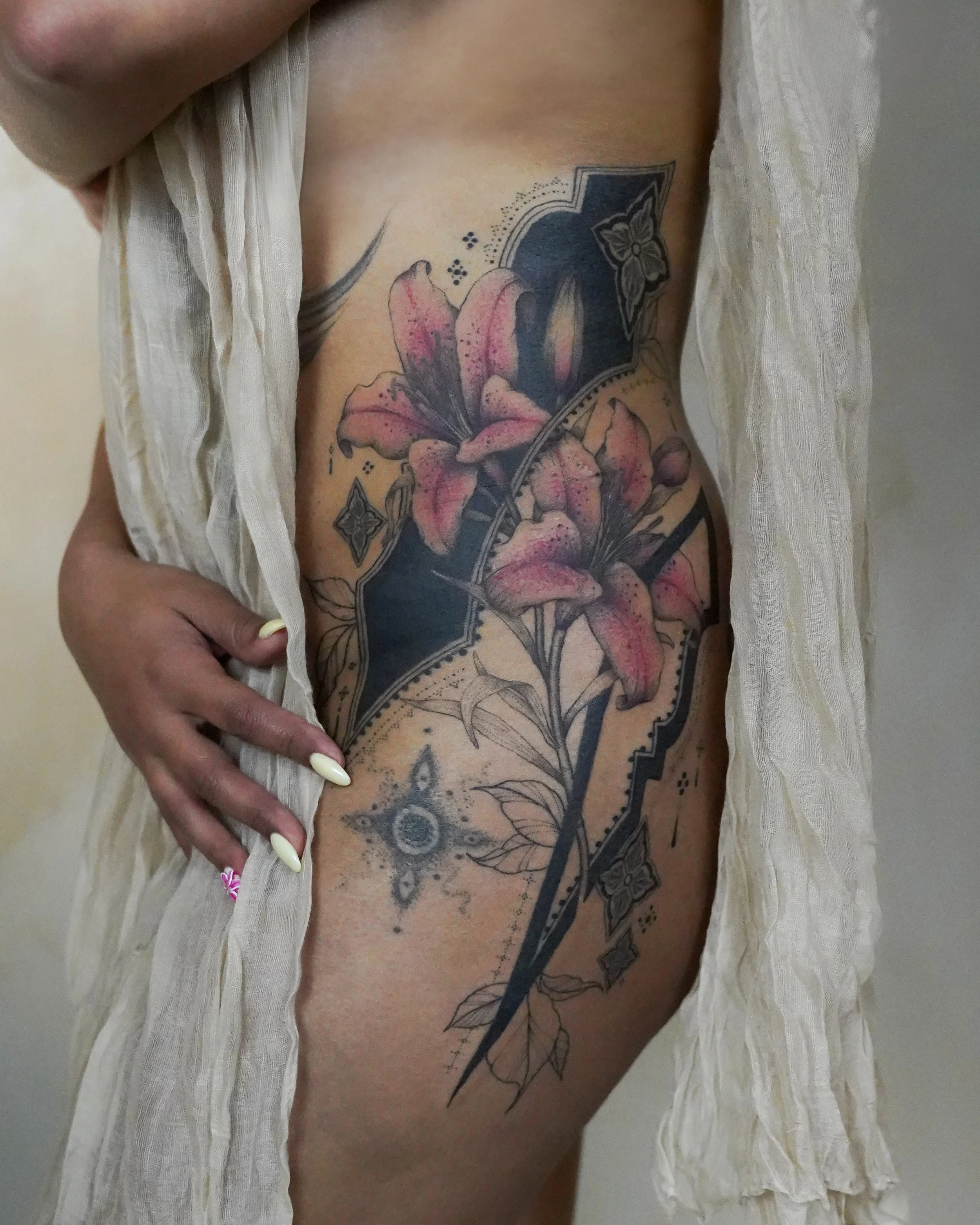 Tattoo of pink lilies with black and gray decorative elements on the person's side, partially covered by a light-colored fabric.