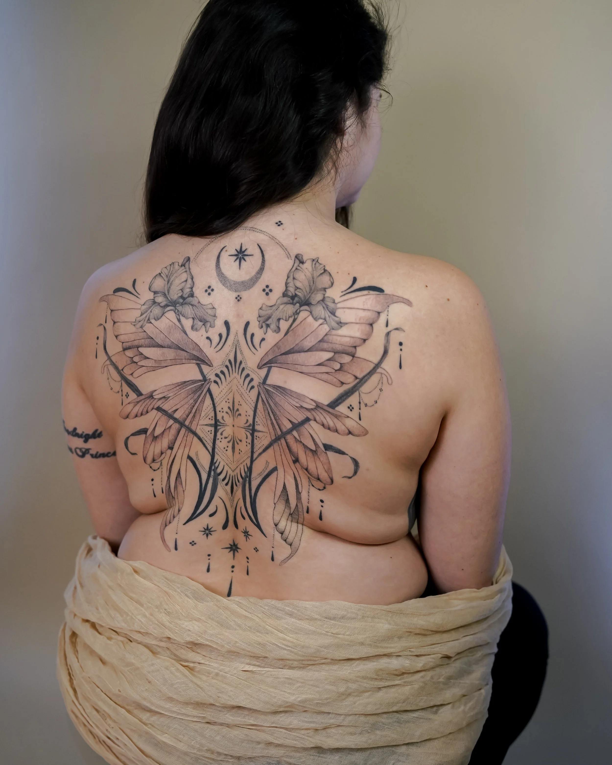A woman with dark hair shows her back tattoo featuring butterflies, moons, stars, and decorative patterns.